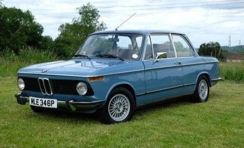 A vintage blue BMW 2002 car parked on a grassy field with trees in the background.