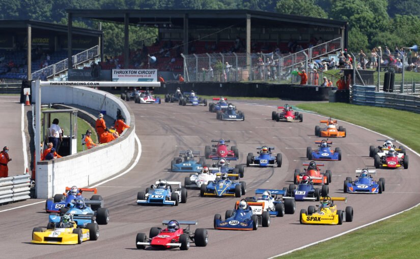 Vintage racing cars on a race track during a race, with spectators in the stands and race officials in orange jumpsuits.