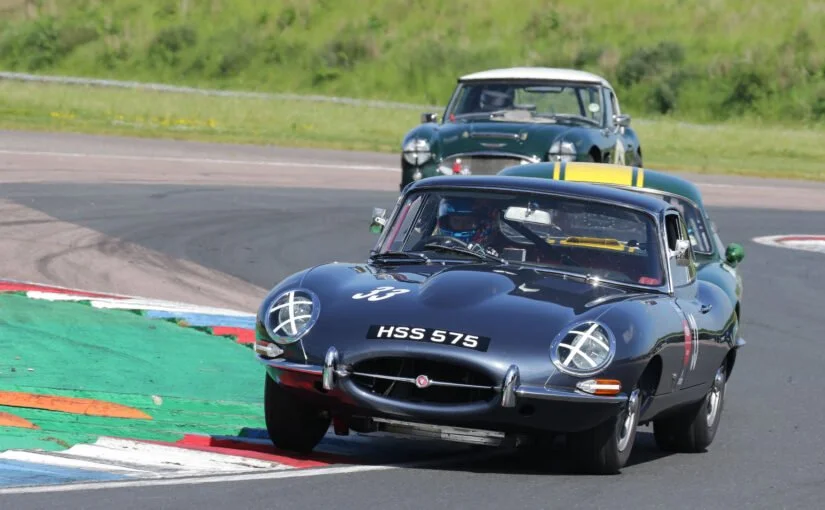 Three vintage race cars driving on a race track. The front car is a black Jaguar with the license plate HSS 575, followed by a silver car and another dark-colored car in the background.