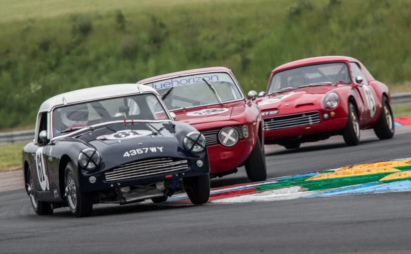 Three vintage racing cars on a race track navigating a turn, with grassy area in the background.