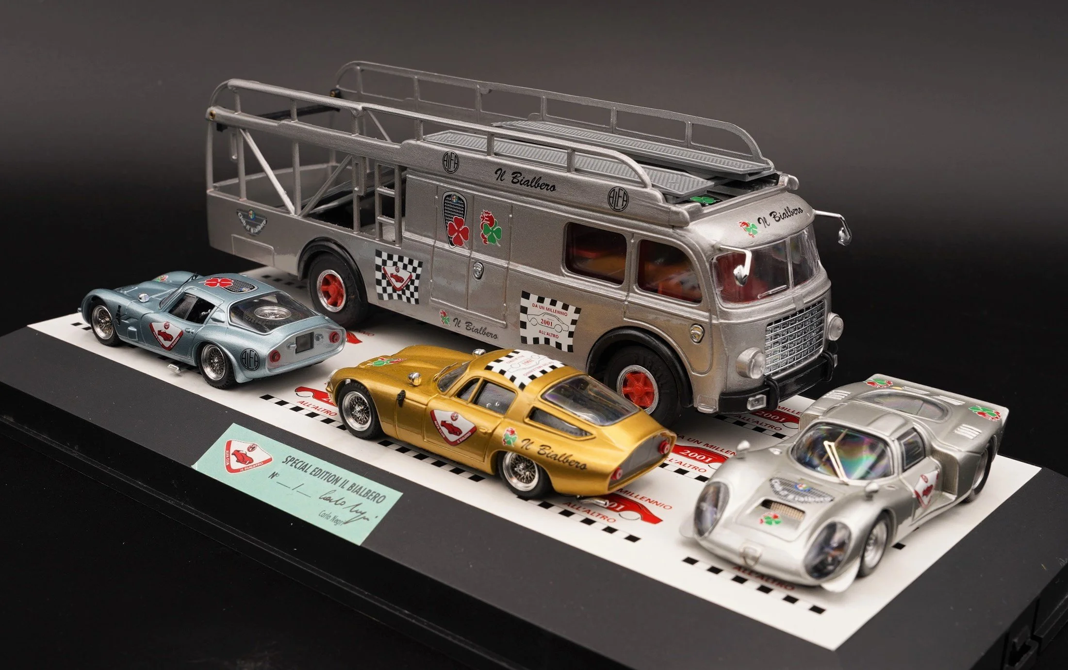 A model set featuring a small silver vintage bus with red hubcaps, alongside three miniature racing cars in silver, gold, and white, all decorated with racing decals and logos, displayed on a black platform with red and white labels.