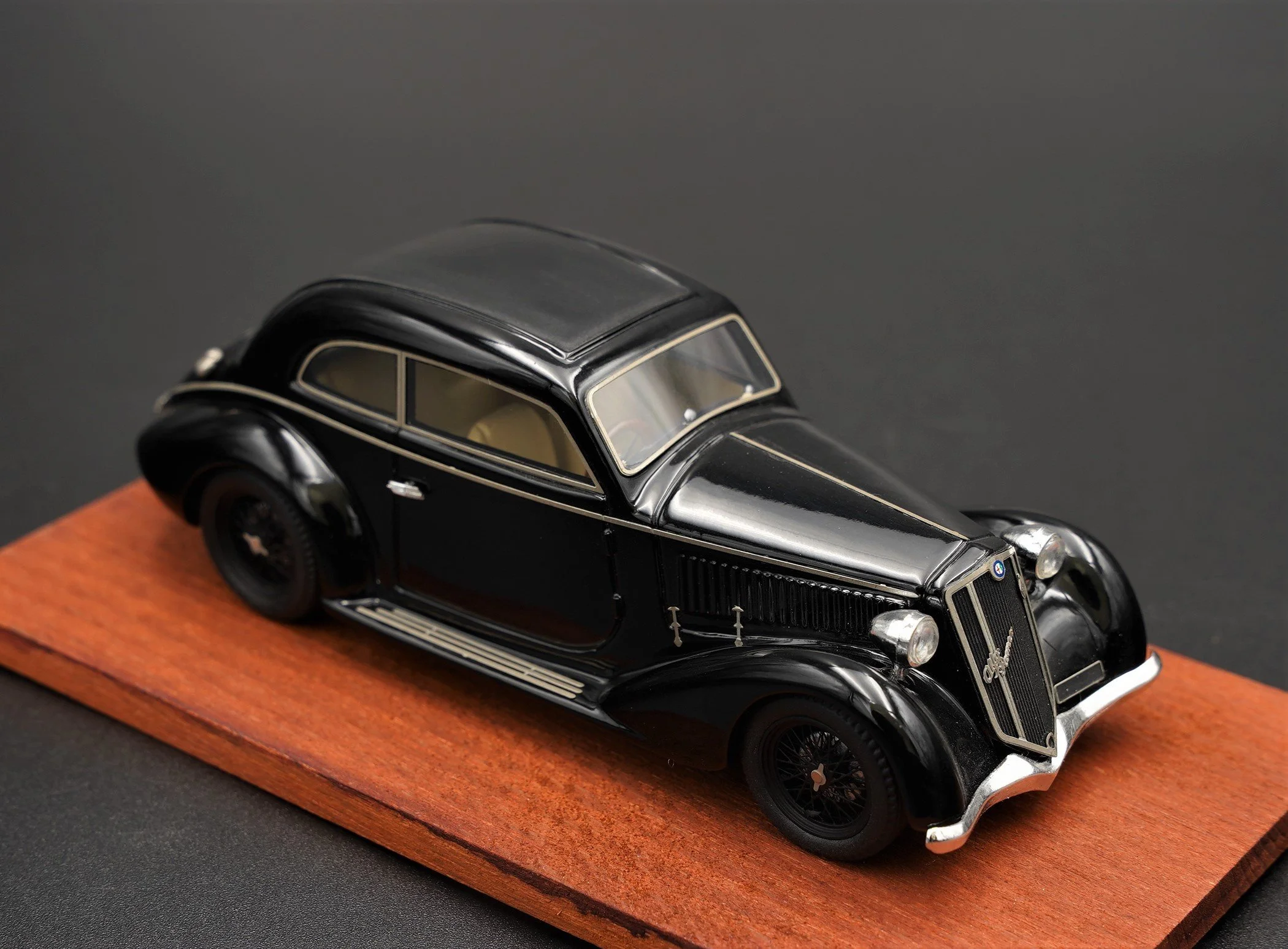 A vintage black model car with intricate detailing, mounted on a wooden base, viewed from above.