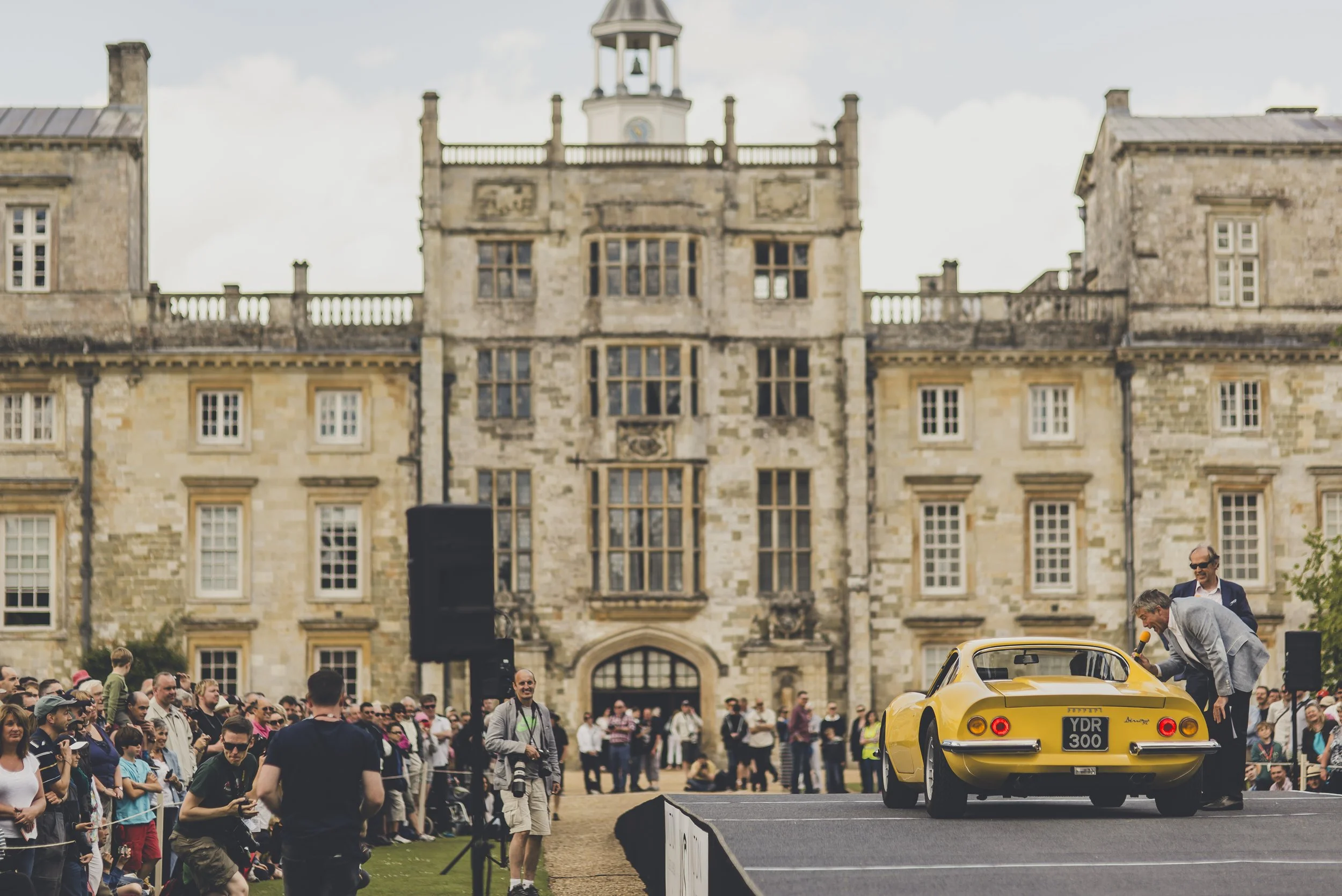 A yellow vintage sports car on display at an outdoor event in front of a historic stone building with a crowd of people watching.