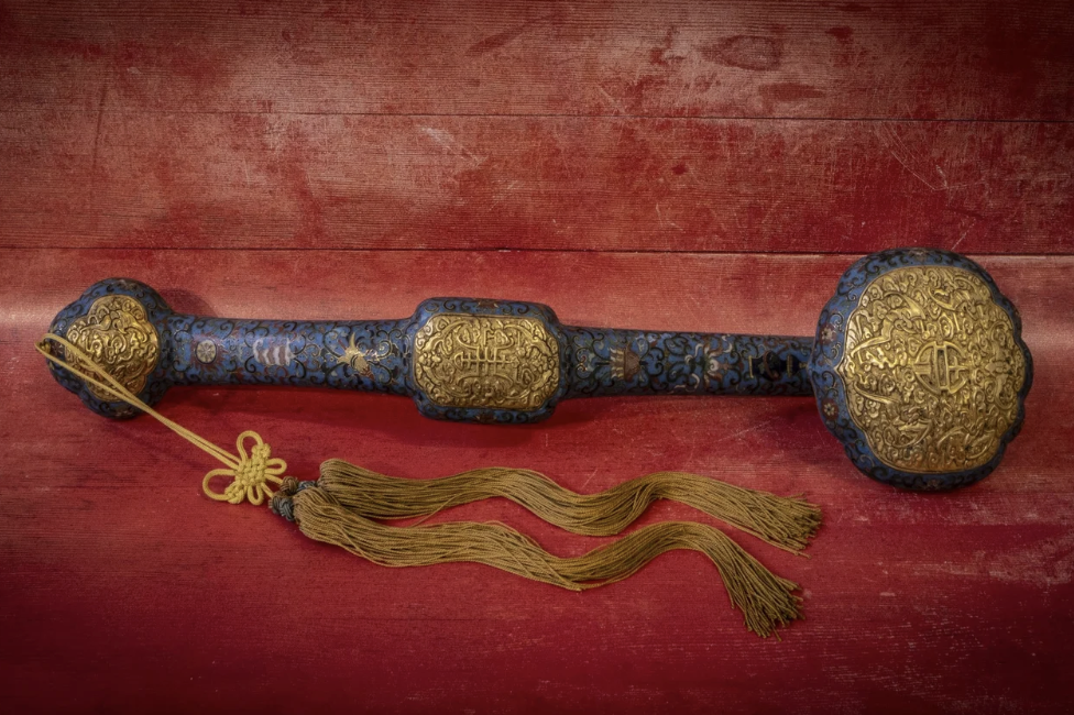Ornate ceremonial staff or scepter with intricate gold engravings and blue embellishments, featuring tassels hanging from its handle, laid on a red surface.