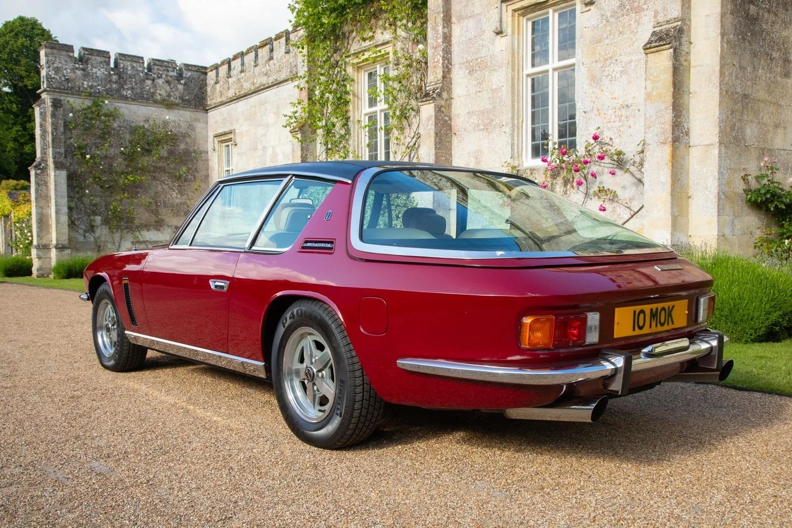 A vintage red sports car parked on a gravel driveway in front of a stone mansion with climbing roses and windows.