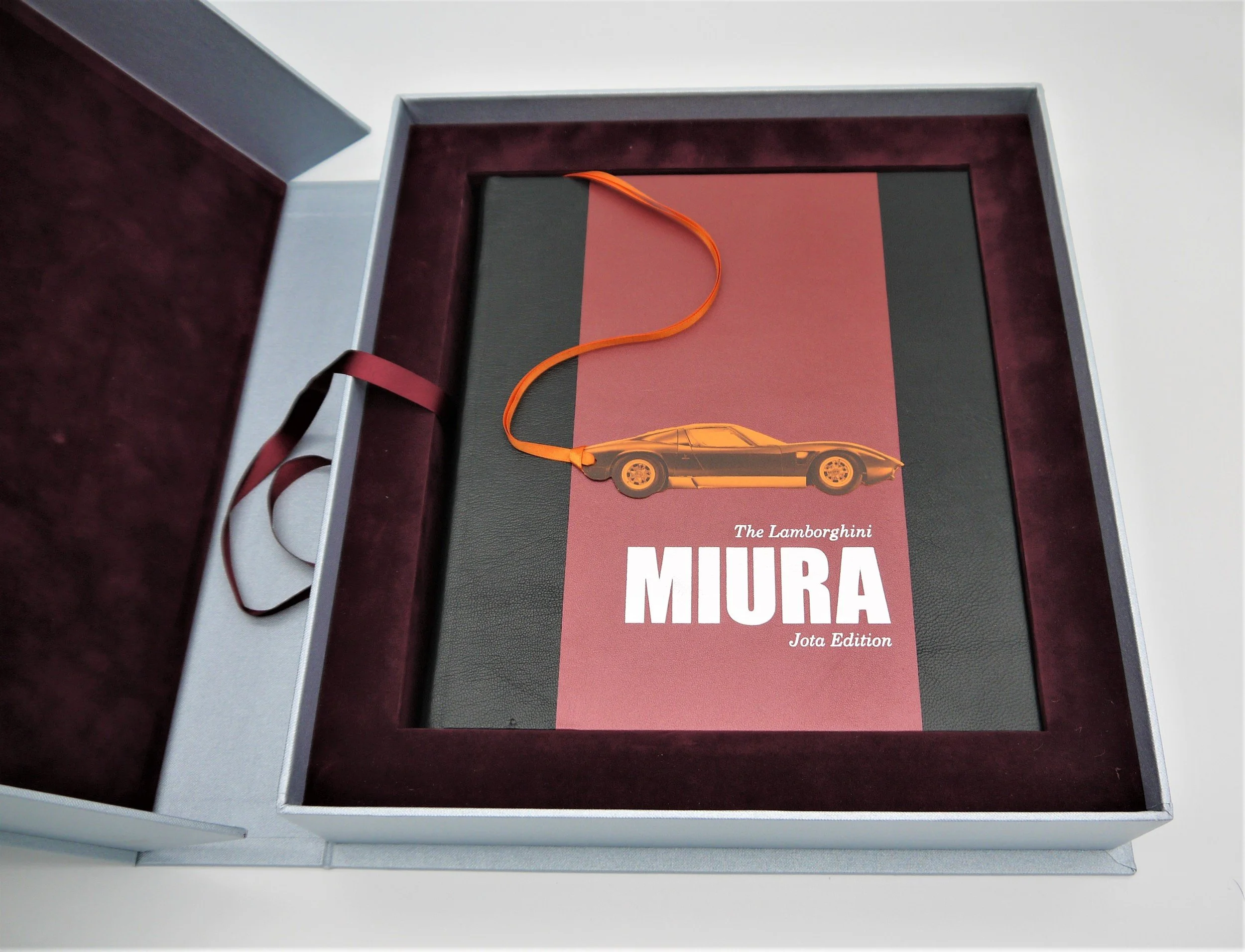 An open luxury presentation box with a book titled 'The Lamborghini MIURA Jota Edition,' featuring a black and red cover with an orange ribbon and a small image of a Lamborghini Miura car.