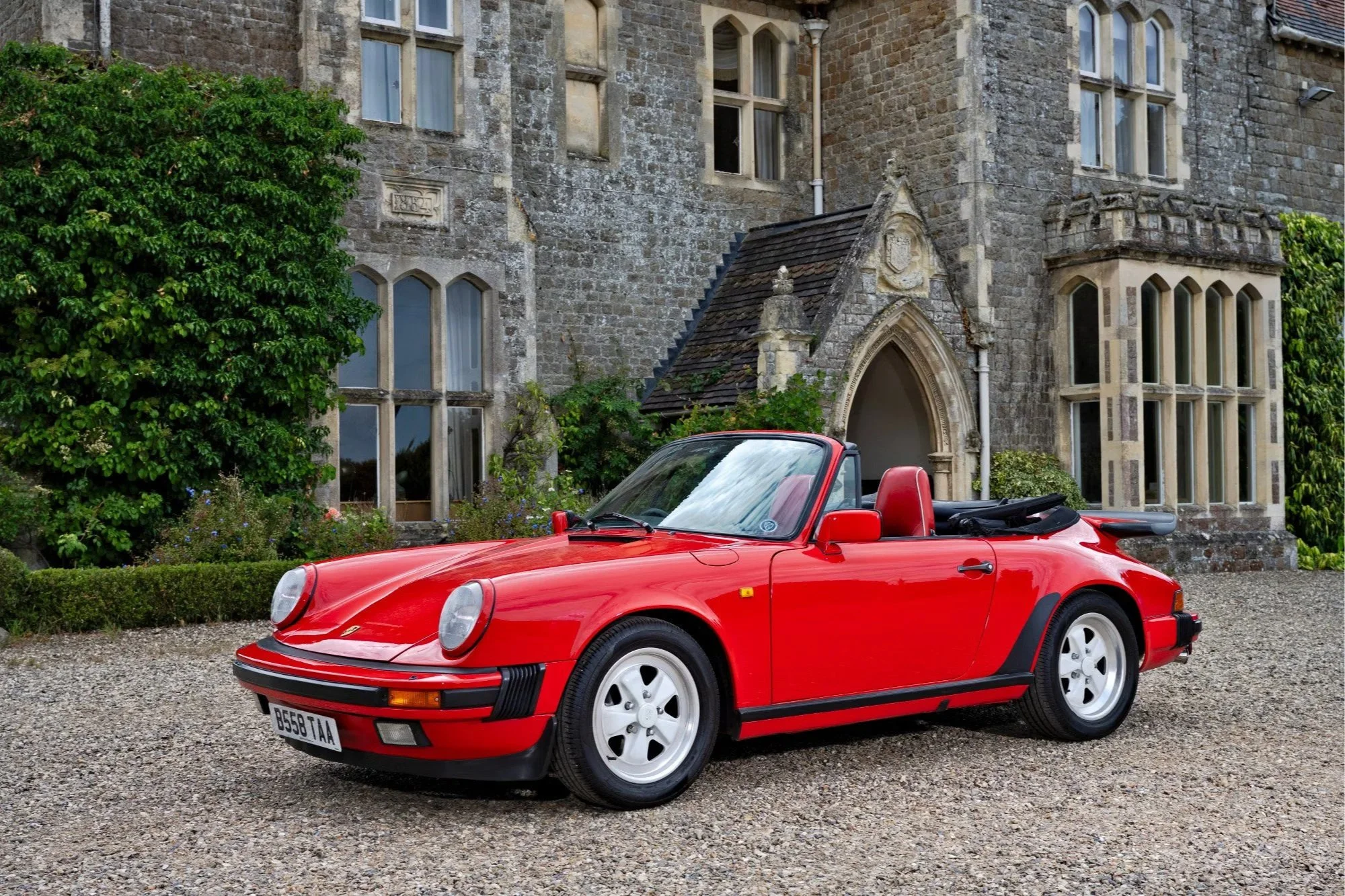 A red Porsche convertible parked outside a stone building with arched windows and greenery.