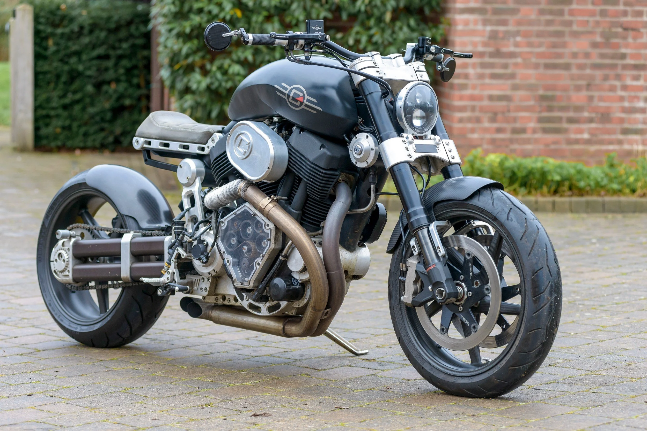 A custom motorcycle parked on a paved driveway with a brick wall and green plants in the background.