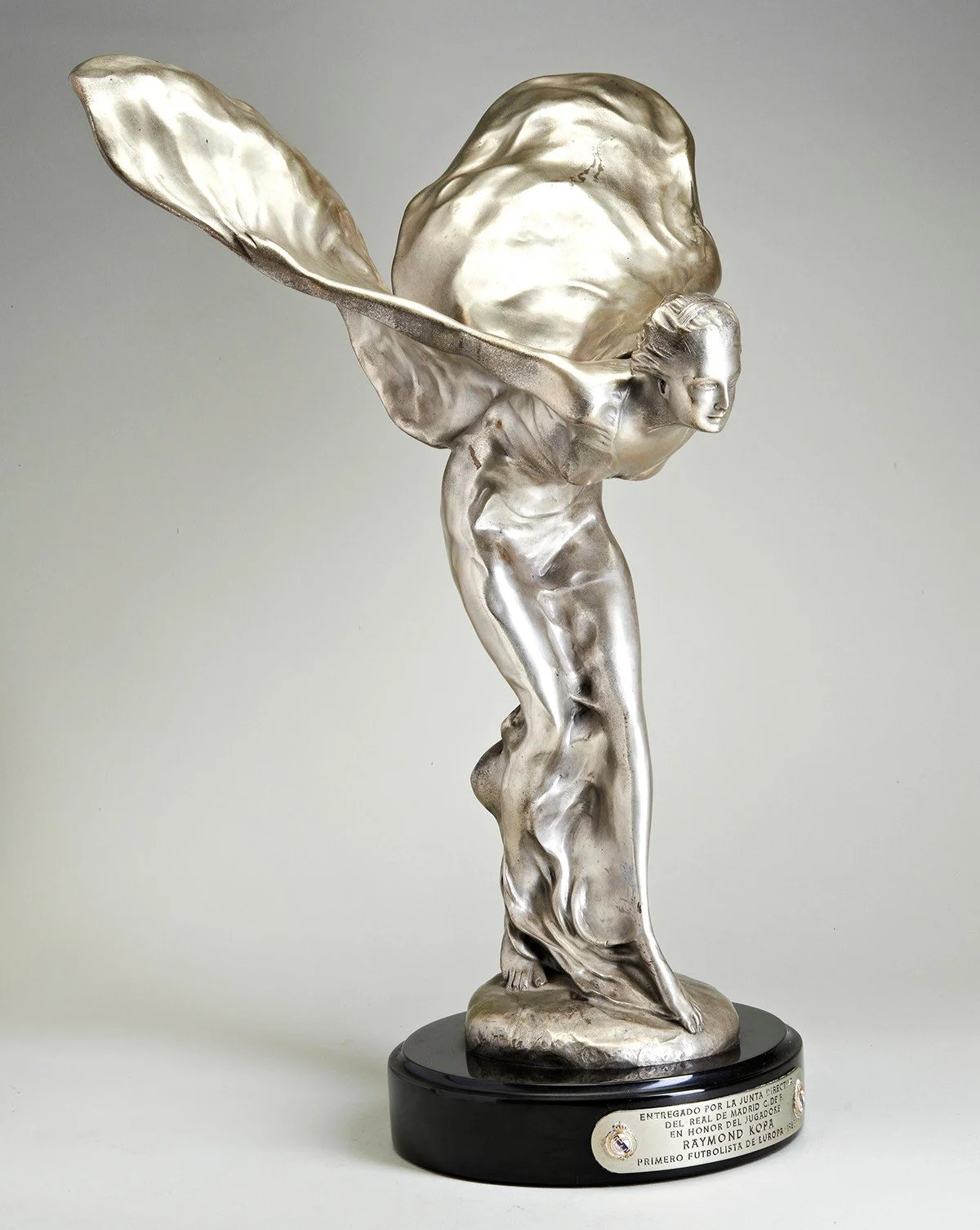 A silver-colored sculpture of a woman with wings, in a dynamic pose, mounted on a black base.