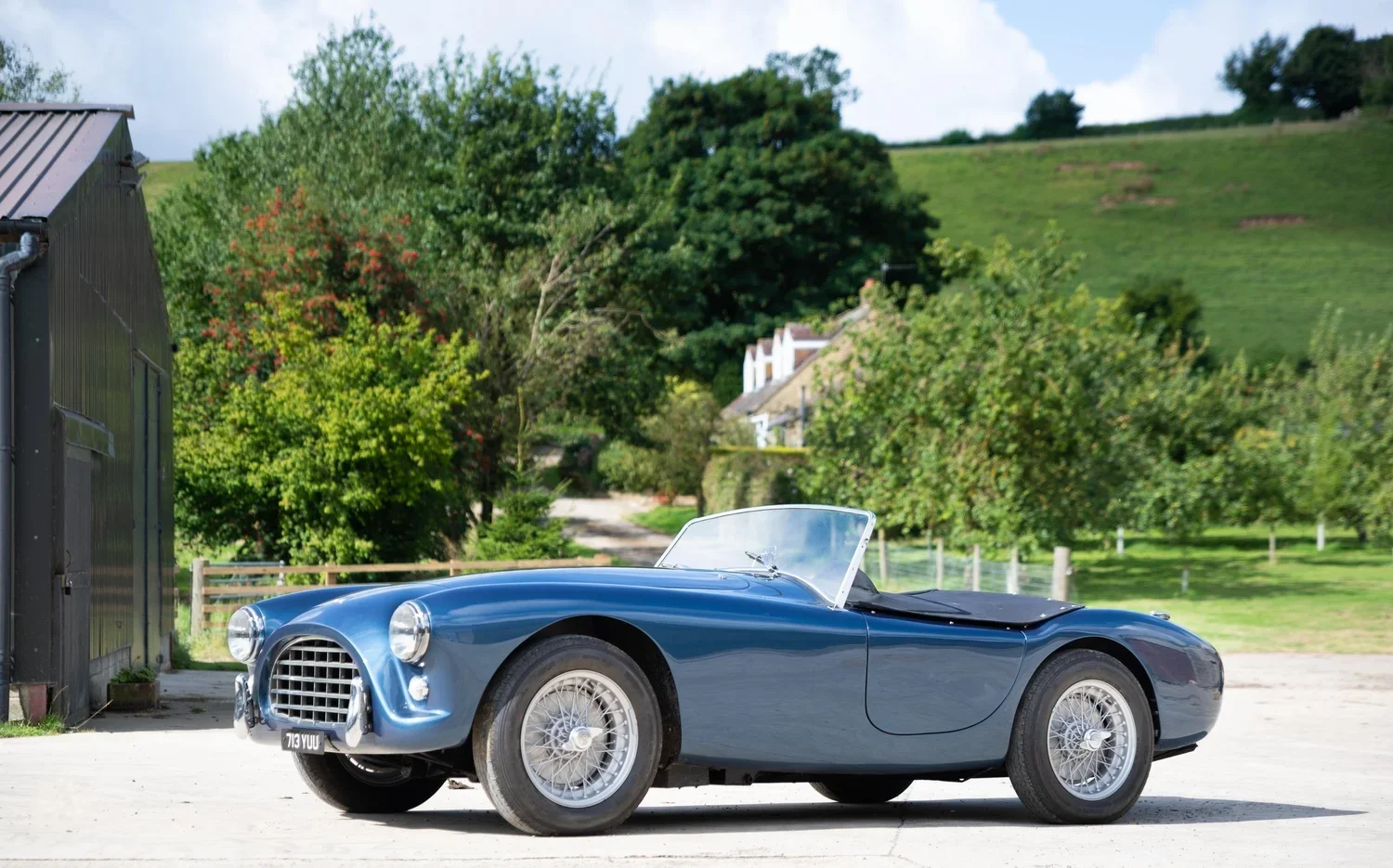 A vintage blue convertible sports car parked outdoors on a concrete surface, with lush green trees, hills, and a few houses in the background.