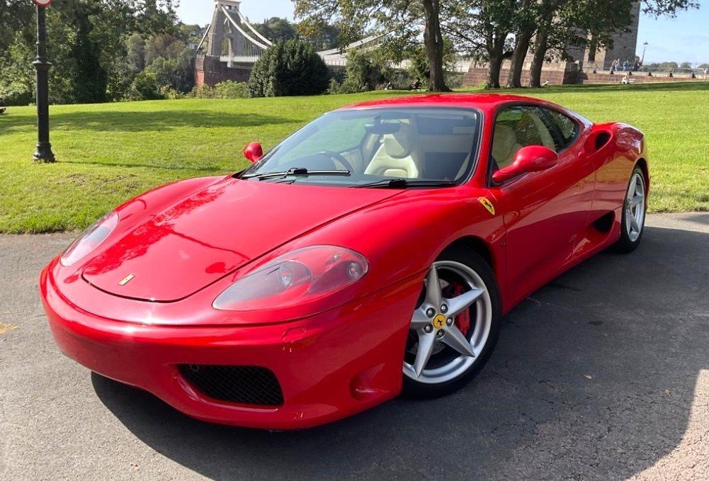 A red Ferrari sports car parked on a pavement near a grassy area with trees and a bridge in the background.