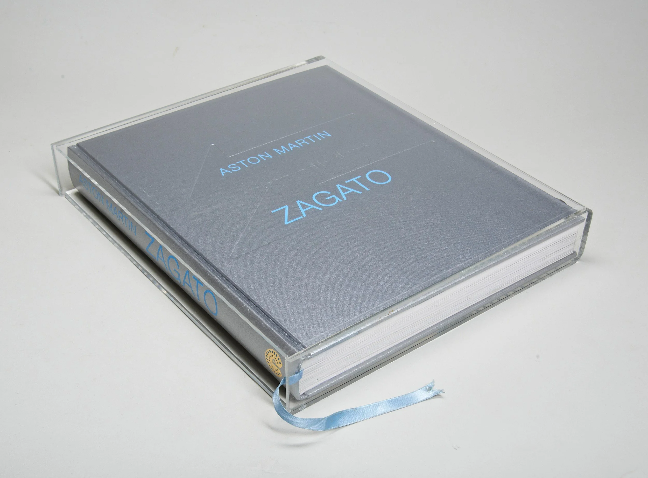 A book titled 'Zagato' by Aston Martin, encased in a clear plastic protective cover with a blue ribbon bookmark.