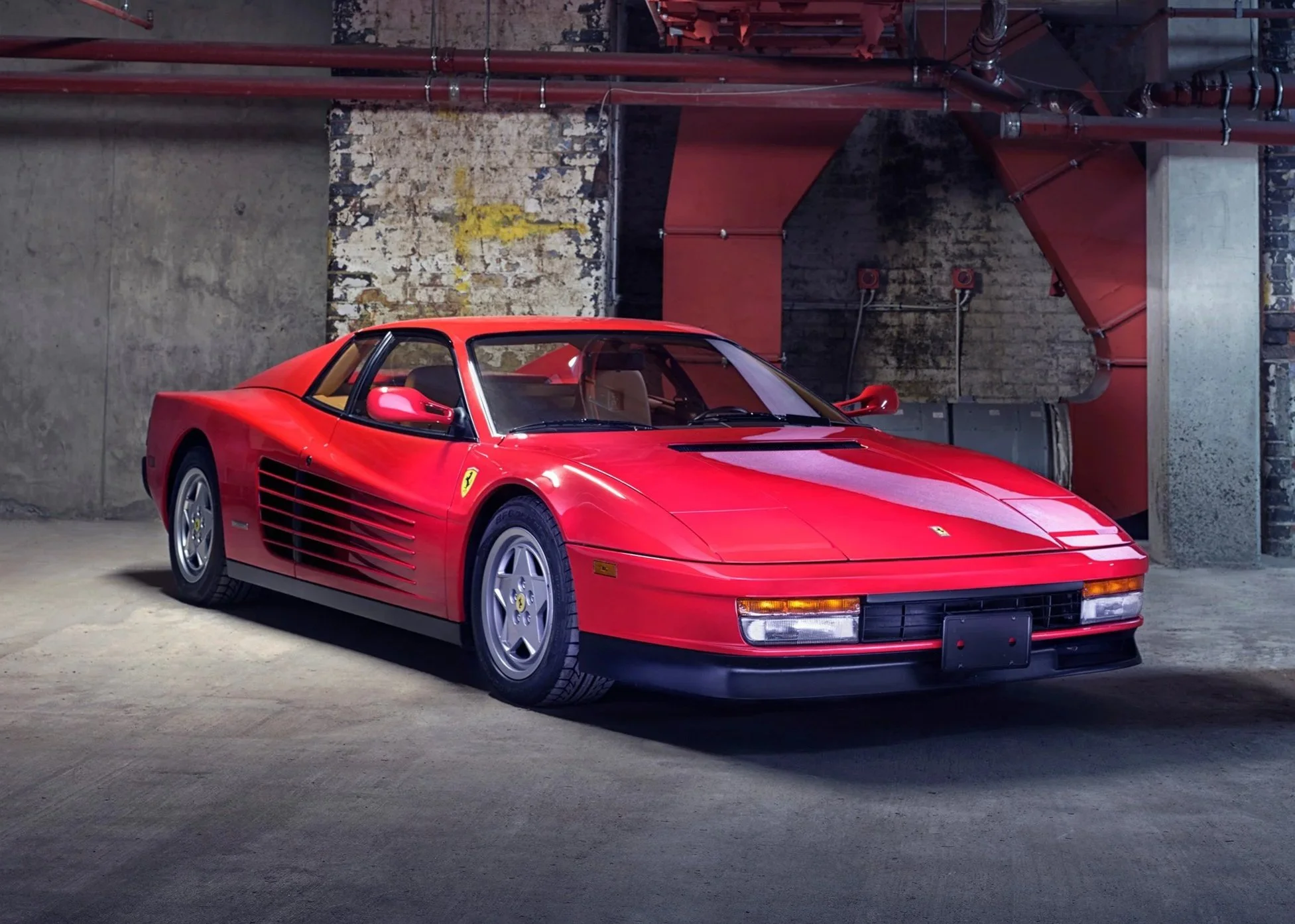 A red Ferrari sports car parked inside an industrial garage with exposed brick walls and pipes.