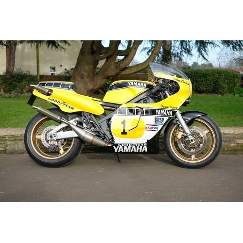Yellow Yamaha racing motorcycle with racing decals parked on the street near a tree and some greenery in the background.