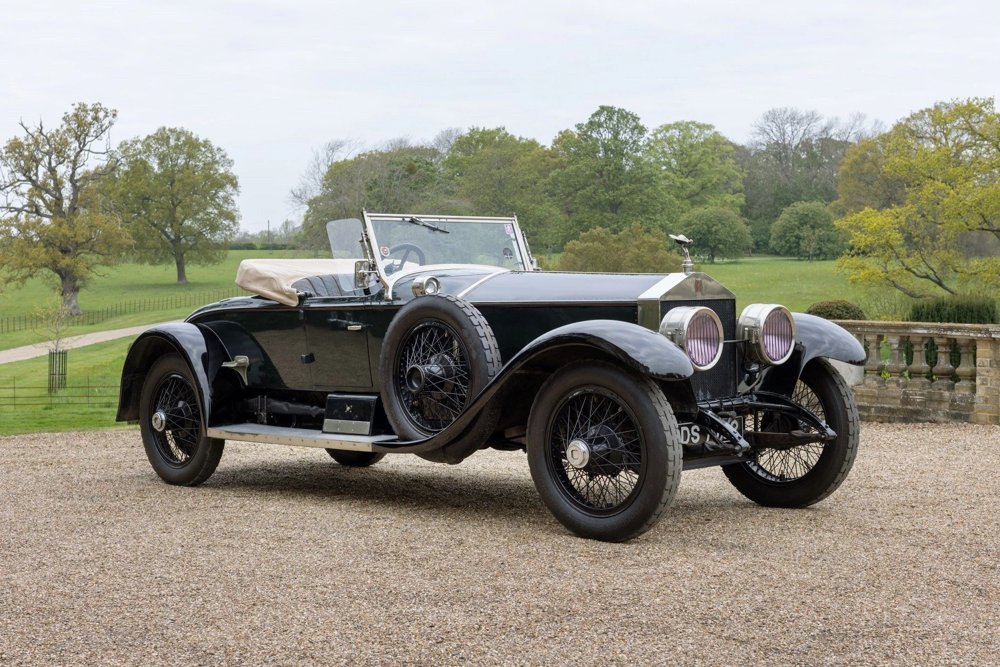A vintage black open-top classic car with wire-spoke wheels parked on a gravel surface with a lush green park and trees in the background.