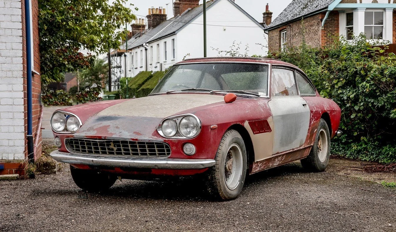 An old, weathered red sports car parked on a driveway next to a house, with signs of rust and peeling paint.