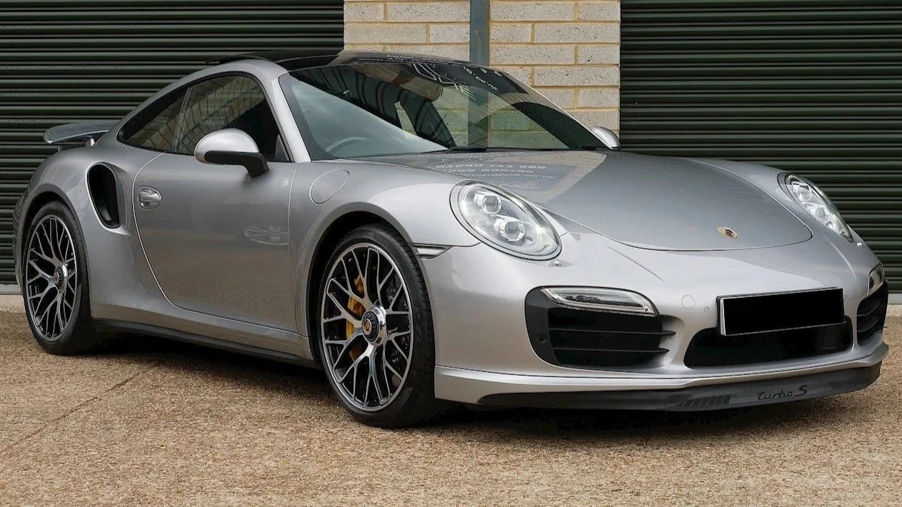Silver Porsche 911 Turbo S sports car parked in front of a brick wall and garage doors.