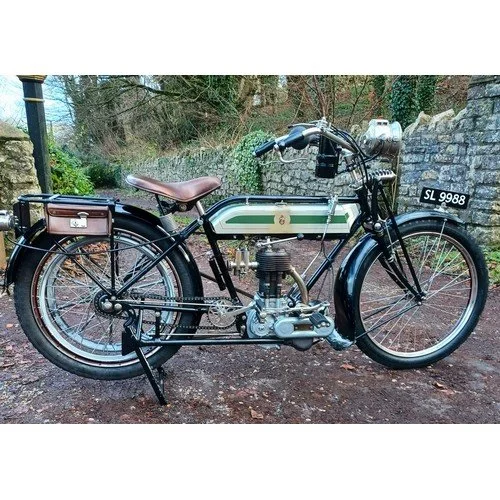 Vintage motorcycle with a black frame, brown leather seat, and a white license plate reading 'SL 9988', parked on a dirt path with trees and a stone wall in the background.