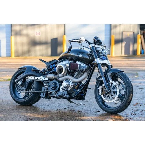 Black custom motorcycle parked on asphalt in front of a warehouse with metal panels and yellow bollards.