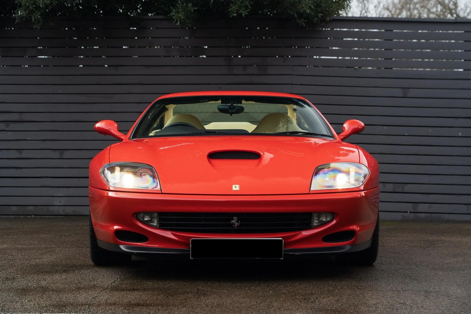 Front view of a red Ferrari sports car parked on a paved surface with a dark horizontal wooden fence in the background.