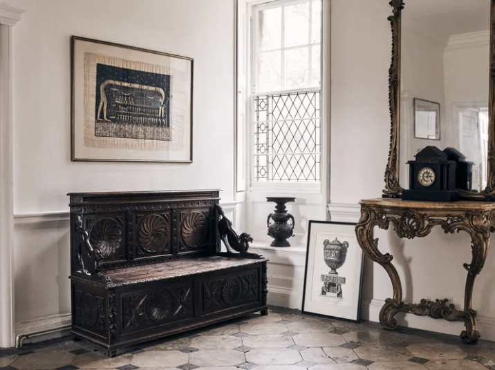 Elegant vintage room with white walls, a black ornate bench, a framed blue artwork, a large window with grid pattern, an antique side table with a clock and a framed art piece on the floor.