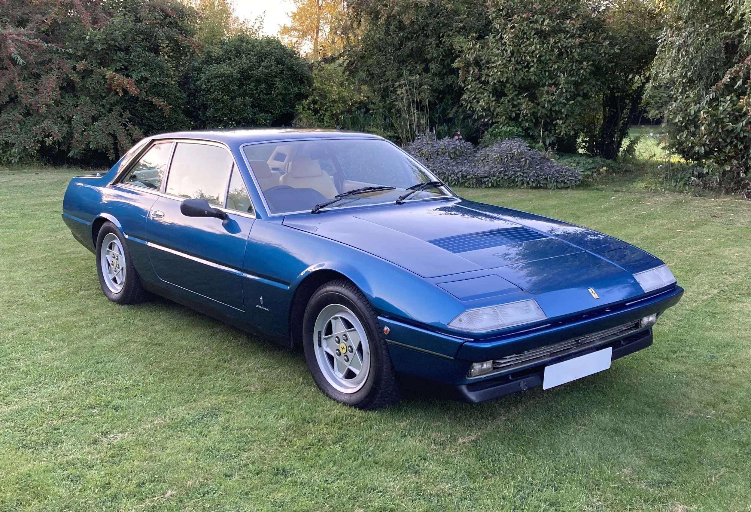 A vintage blue Ferrari sports car parked on a grassy area with trees and bushes in the background.
