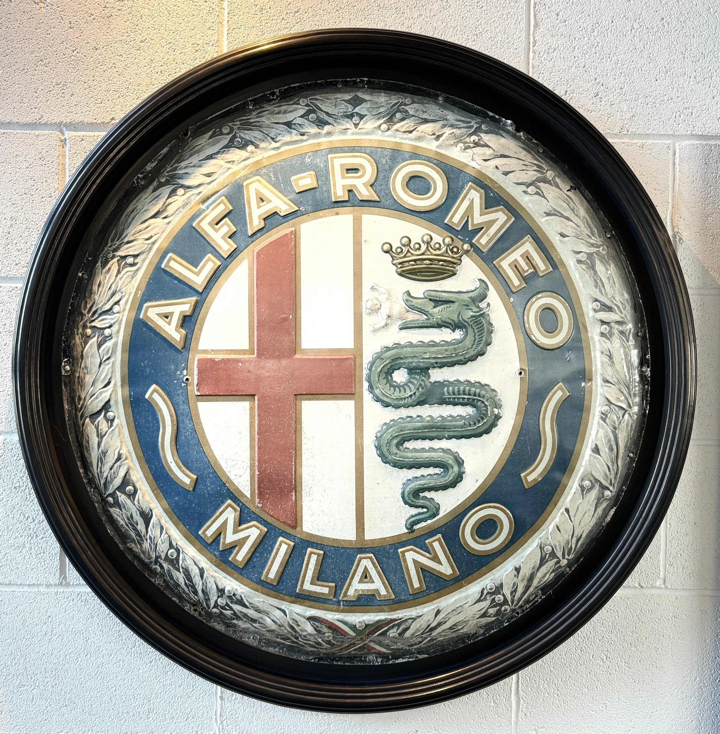 Round sign with a black frame, displaying the Alfa Romeo logo, which includes a red cross on the left, a green snake with a crown on the right, and the text 'ALFA-ROMEO MILANO' around the edge.