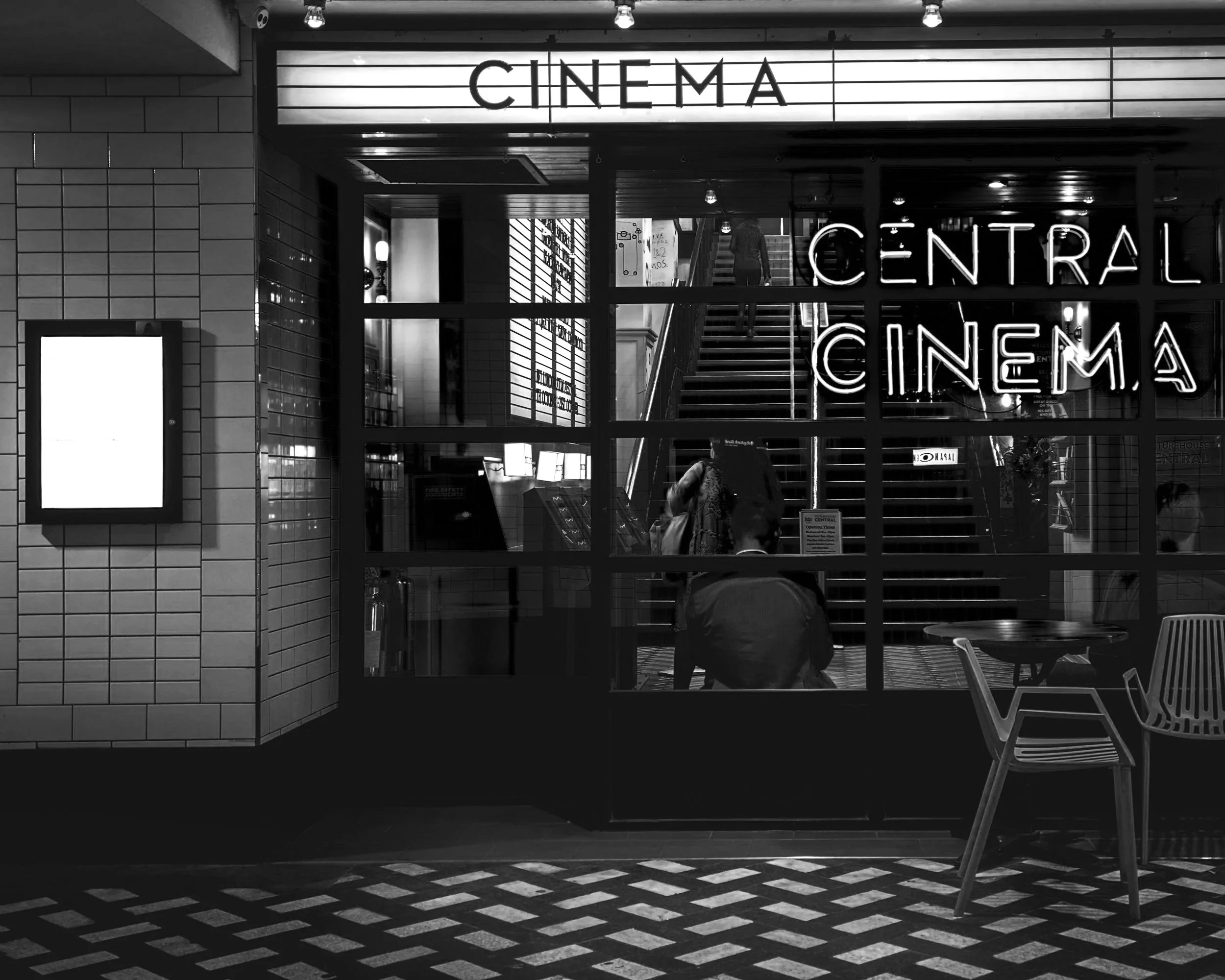 Black and white photo of a cinema entrance with lit signs that say 'CINEMA' and 'CENTRAL CINEMA,' inside visible through glass are people and a staircase.