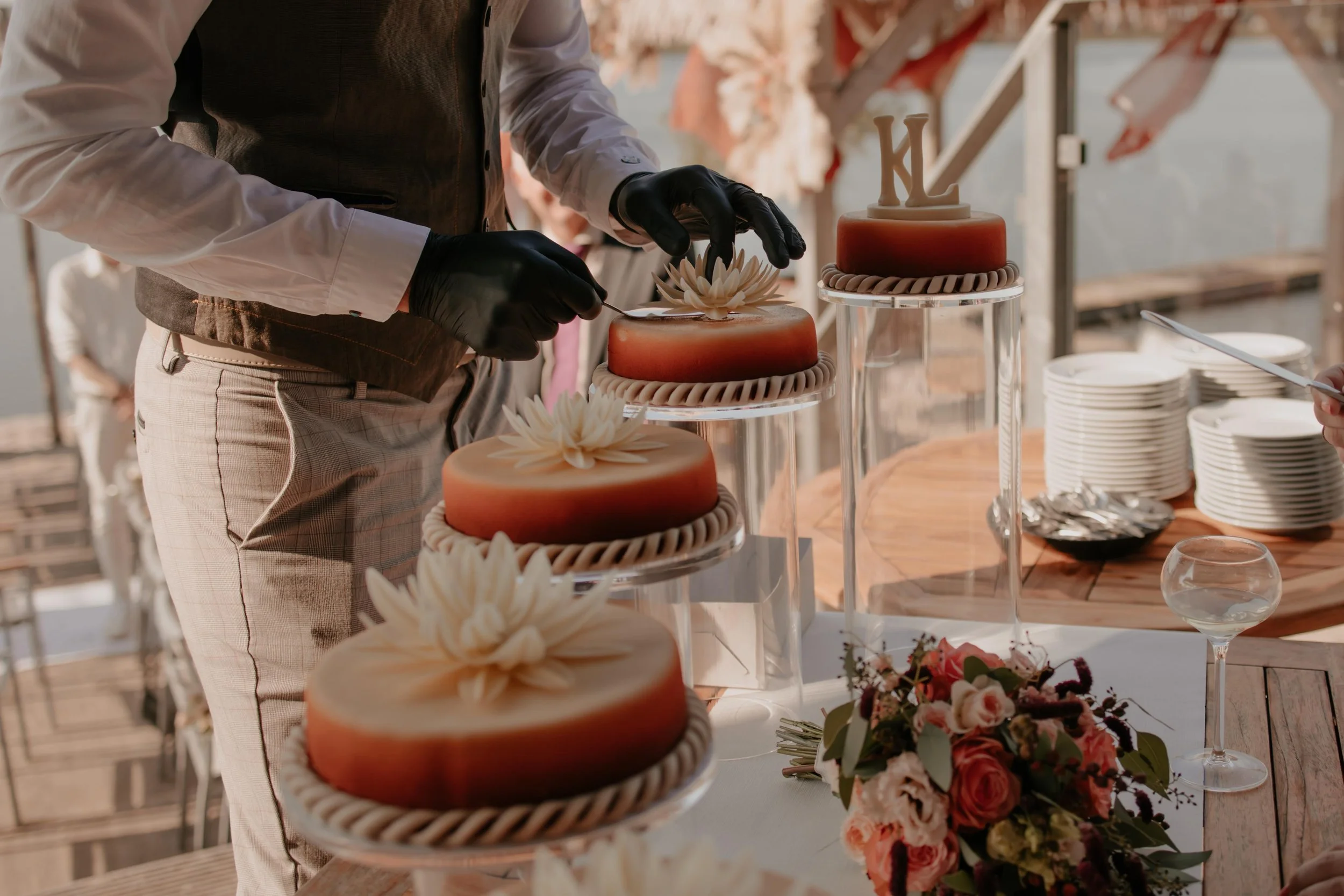 An individual wearing black gloves and a brown vest decorates a tiered cake at a wedding or celebration. The cake features a gradient color design with white floral decorations on top. Several similar cakes are on display, with plates, a bouquet of pink and white flowers, a glass of wine, and a wooden table in the background.