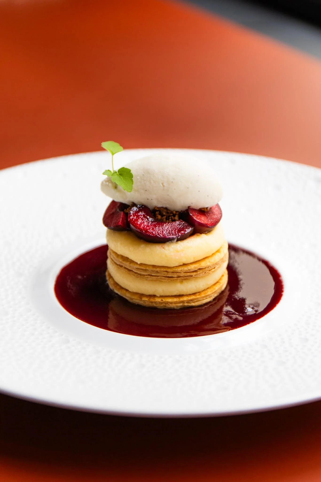 A gourmet dessert consisting of a stack of small pancakes topped with cherries, cream, and garnished with a sprig of herb, served with berry sauce on a white textured plate.