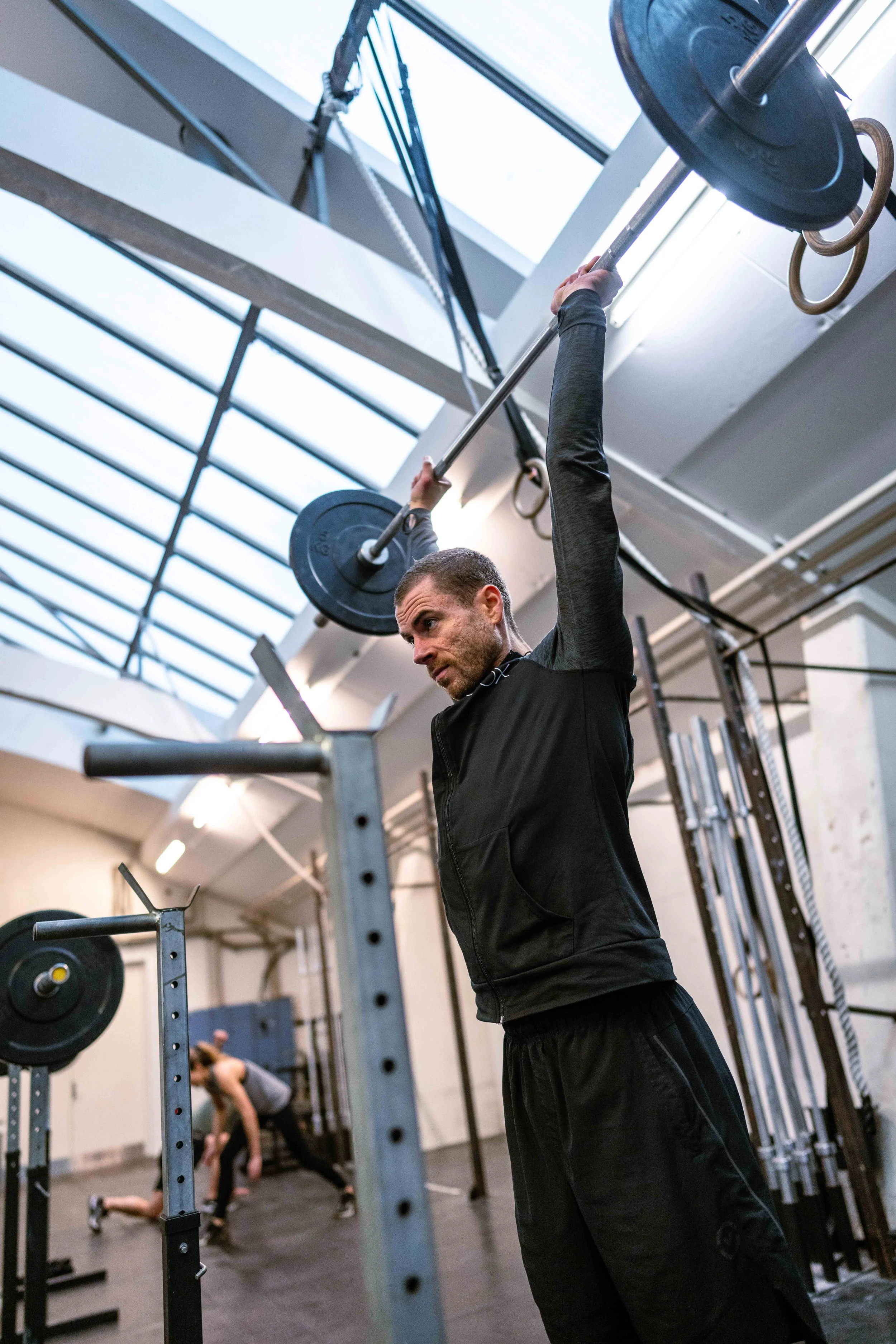 En mand, der løfter en vægtstang over hovedet i et crossfit- eller fitnesscenter, med en anden person i baggrunden, der strækker sig.