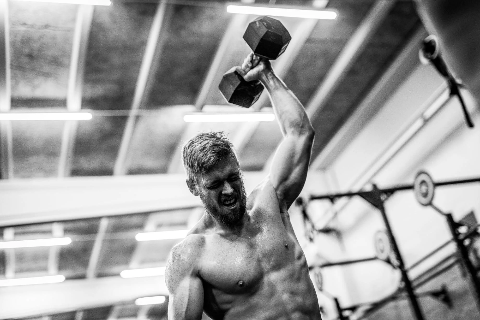 En muskuløs mand med skæg, der har trænet i et fitnesscenter, holder en vægt over hovedet og ser anstrengende ud.