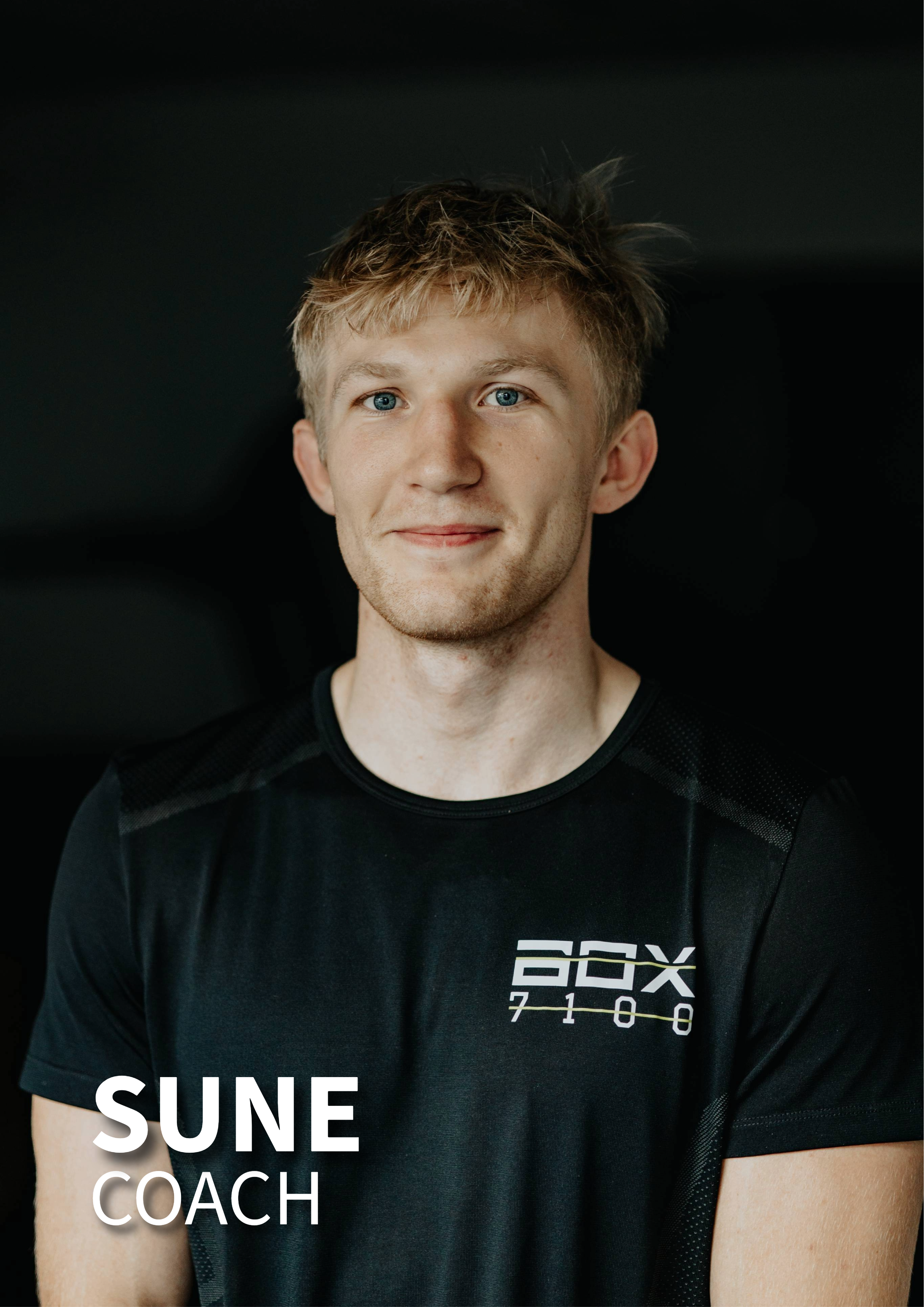 En ung mand med blond, let frygthår, smilende, iført sort trænings T-shirt med tekst og logo, mod mørk baggrund.
