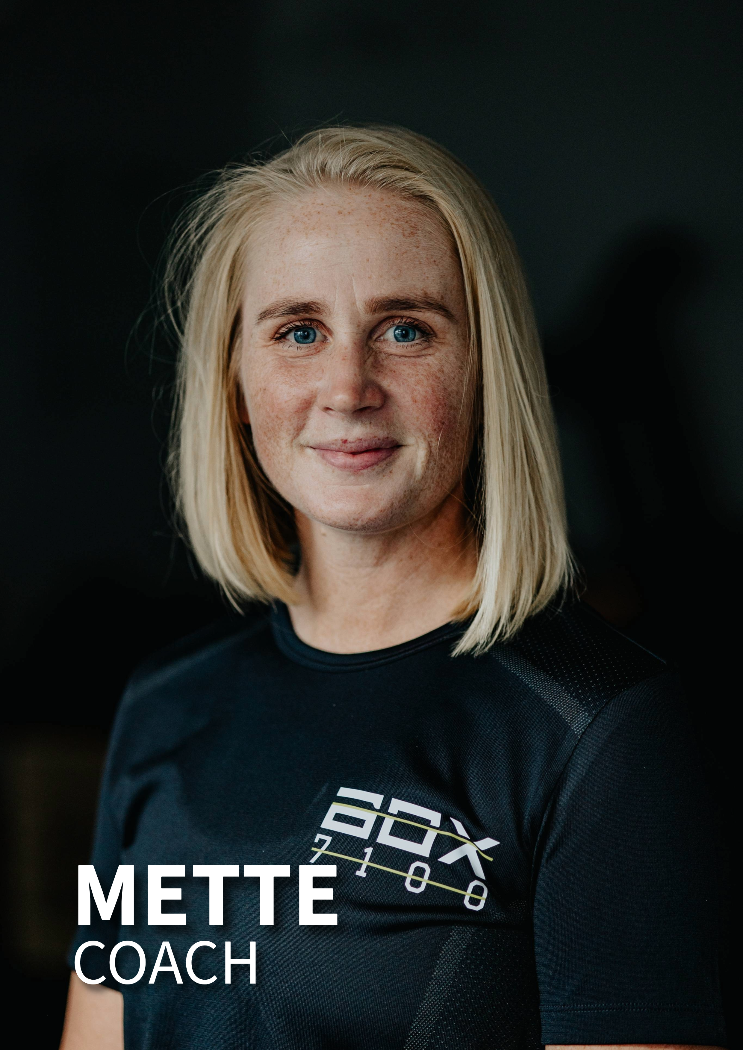 Portræt af en kvinde med blond hår iført sort trøje med logo, stående foran en mørk baggrund.