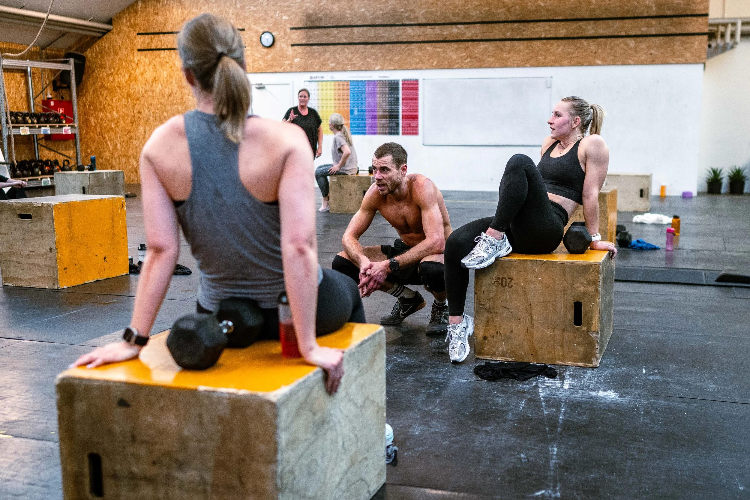 Mennesker, der træner i et crossfit-studie, inklusive to kvinder og en mand, der sidder på træningsbåde, mens en instruktør taler.