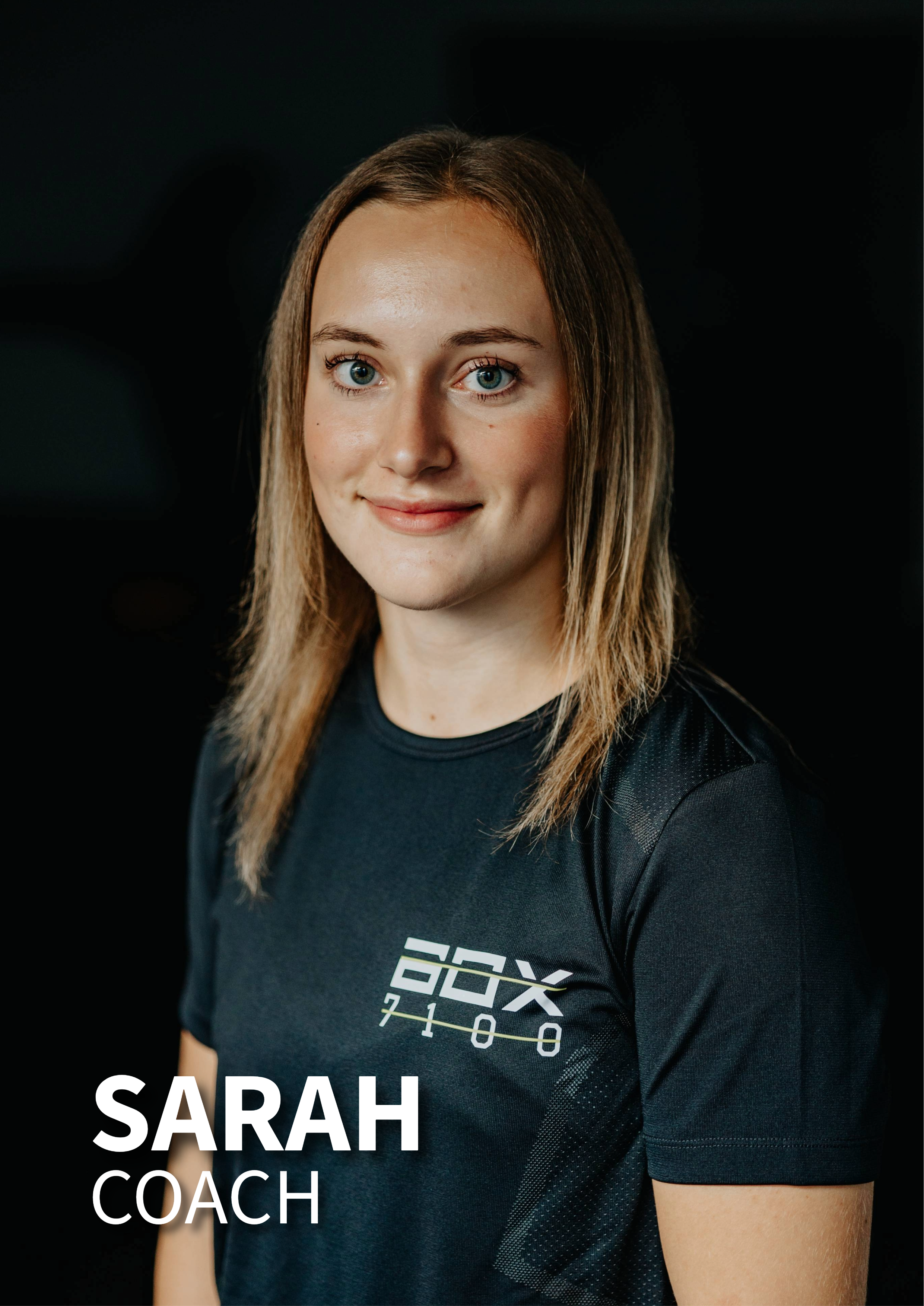 Portræt af en kvinde med navnet Sarah, iført trænings tøj med etiketten 'BOX 7108', mod en sort baggrund.