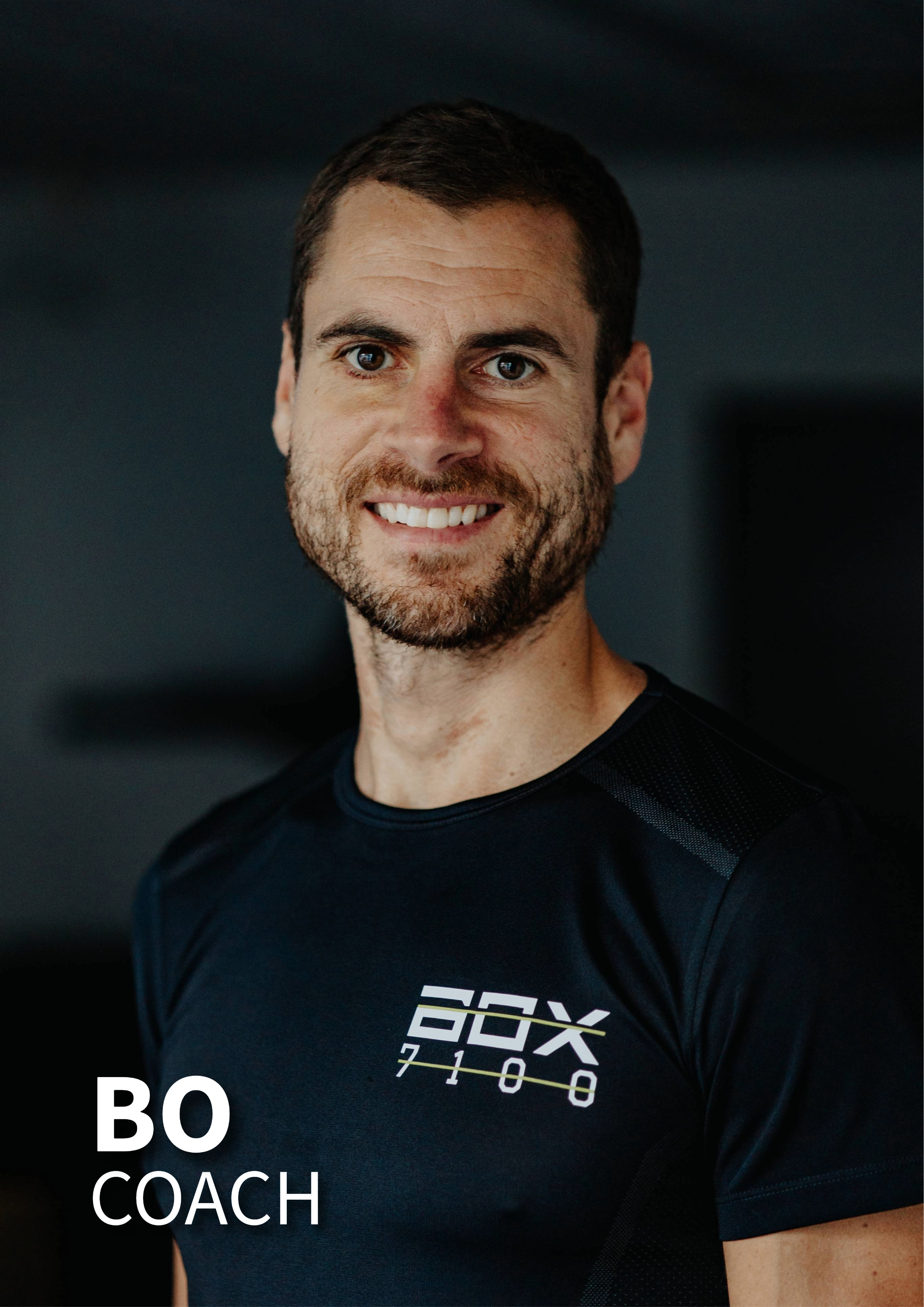 En mand med kort mørkt hår og skæg, smilende, iført sort trænings T-shirt med logoet 'BOXX 7100'.