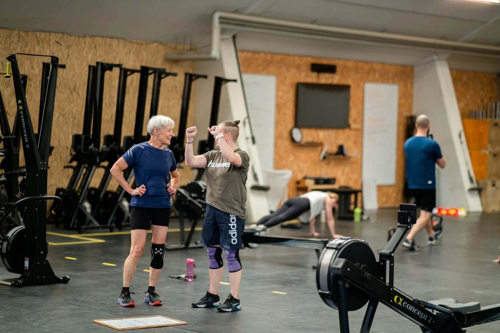To kvinder snakker i et fitnesscenter, en kvinde med kort hvidt hår og en yngre kvinde, begge i træningstøj, mens de smiler og hilser. Der er andre personer i baggrunden, der træner.