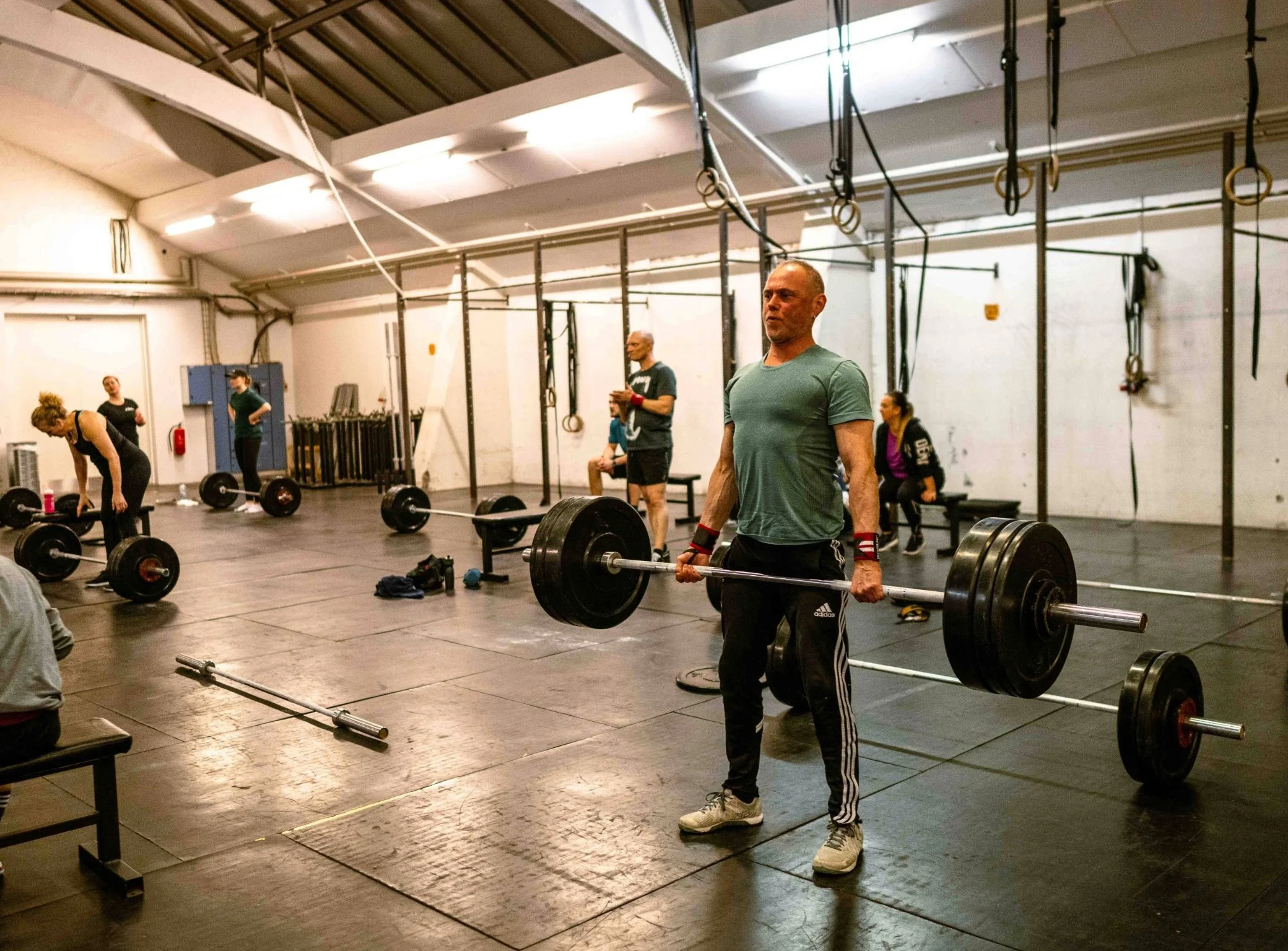 Mennesker træner vægtløftning i et fitnesscenter. En mand i forgrunden løfter en vægtstang med store vægtskiver. Flere personer træner i baggrunden, nogle med vægte og andre sidder på bænke.