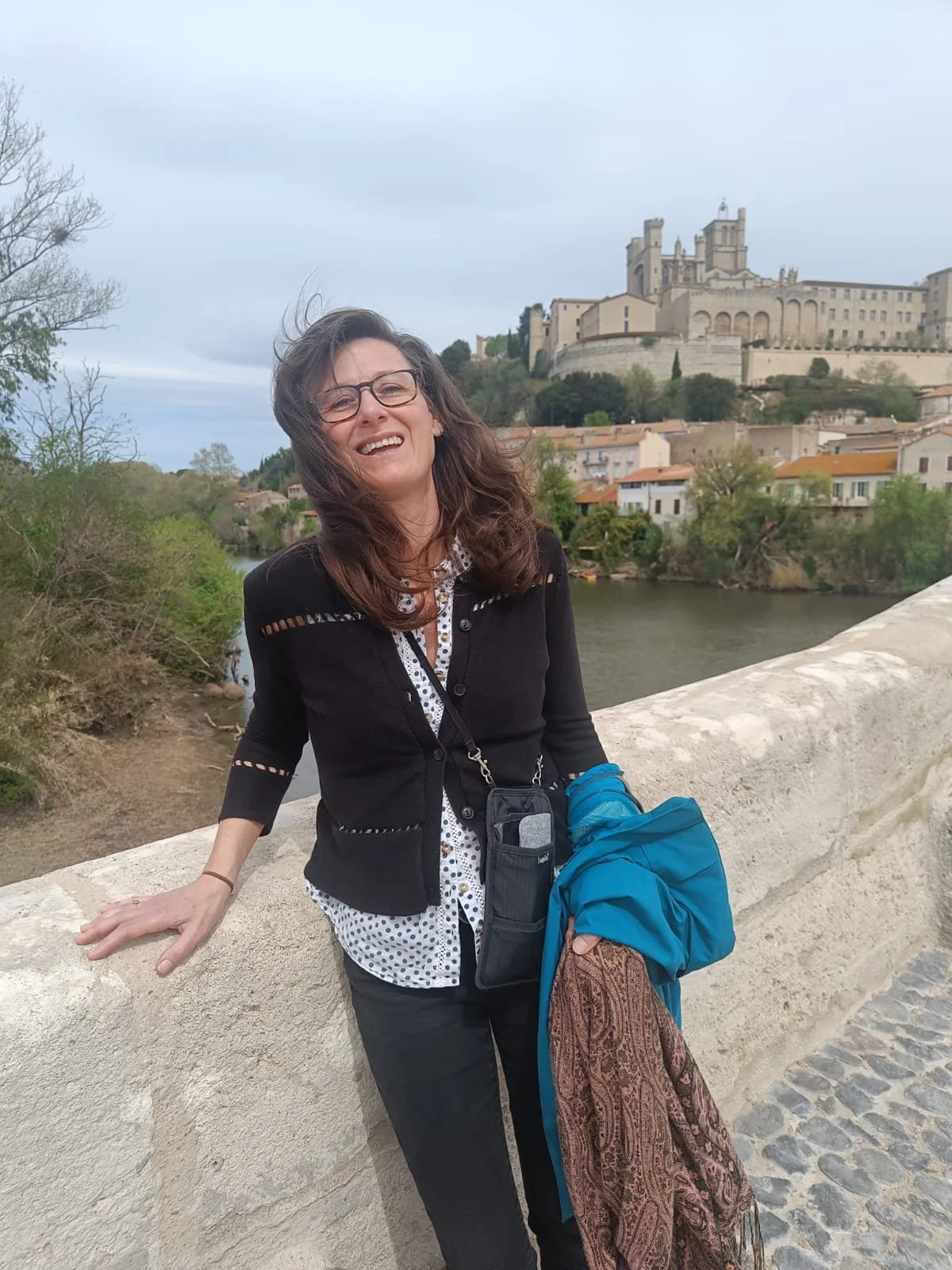 A woman with curly brown hair, wearing glasses, a black cardigan, and dark pants, smiling while standing on a bridge with a historic castle on a hill and river in the background.