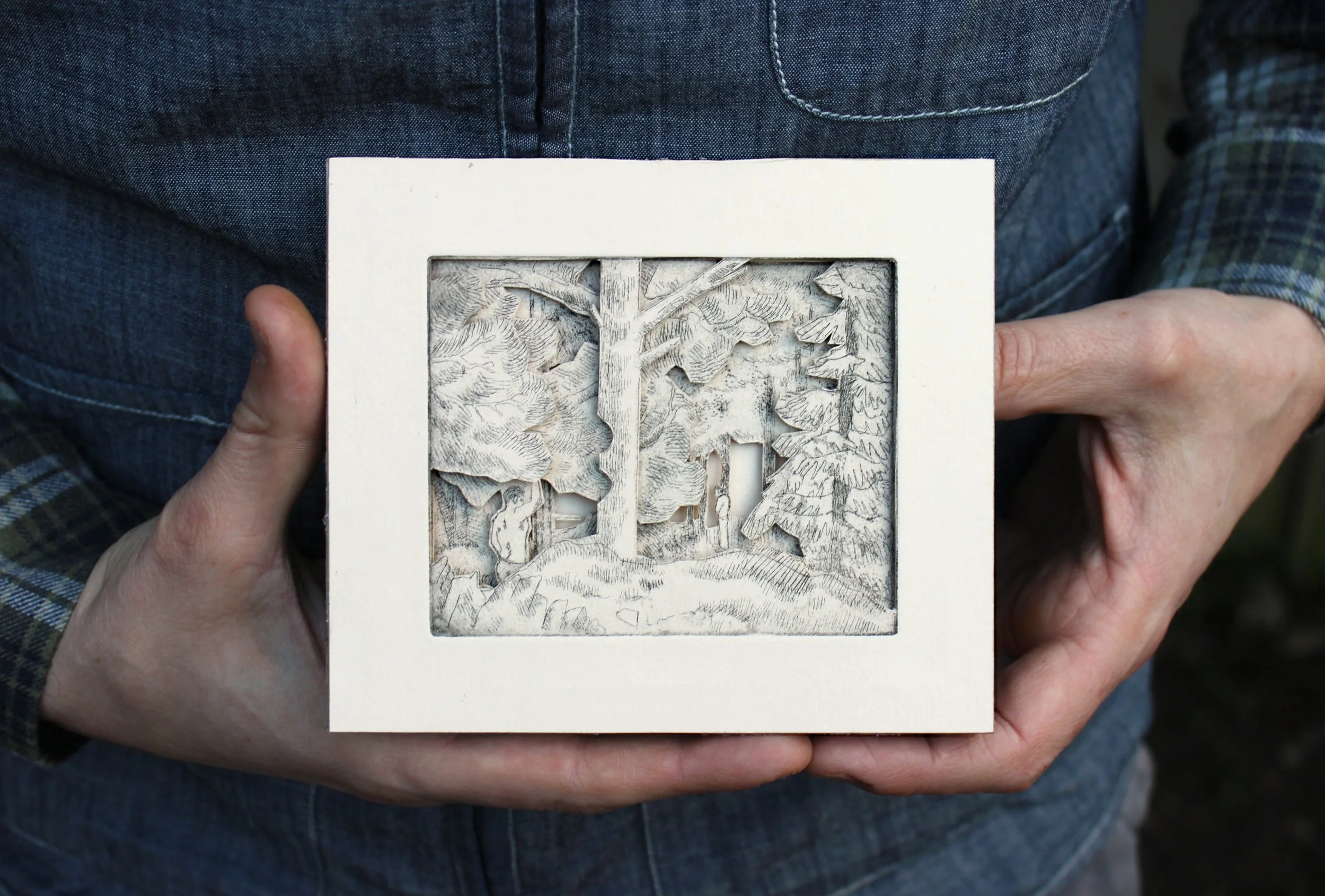 Person holding a white frame with a detailed wood carving of a forest scene, including trees and a small building.