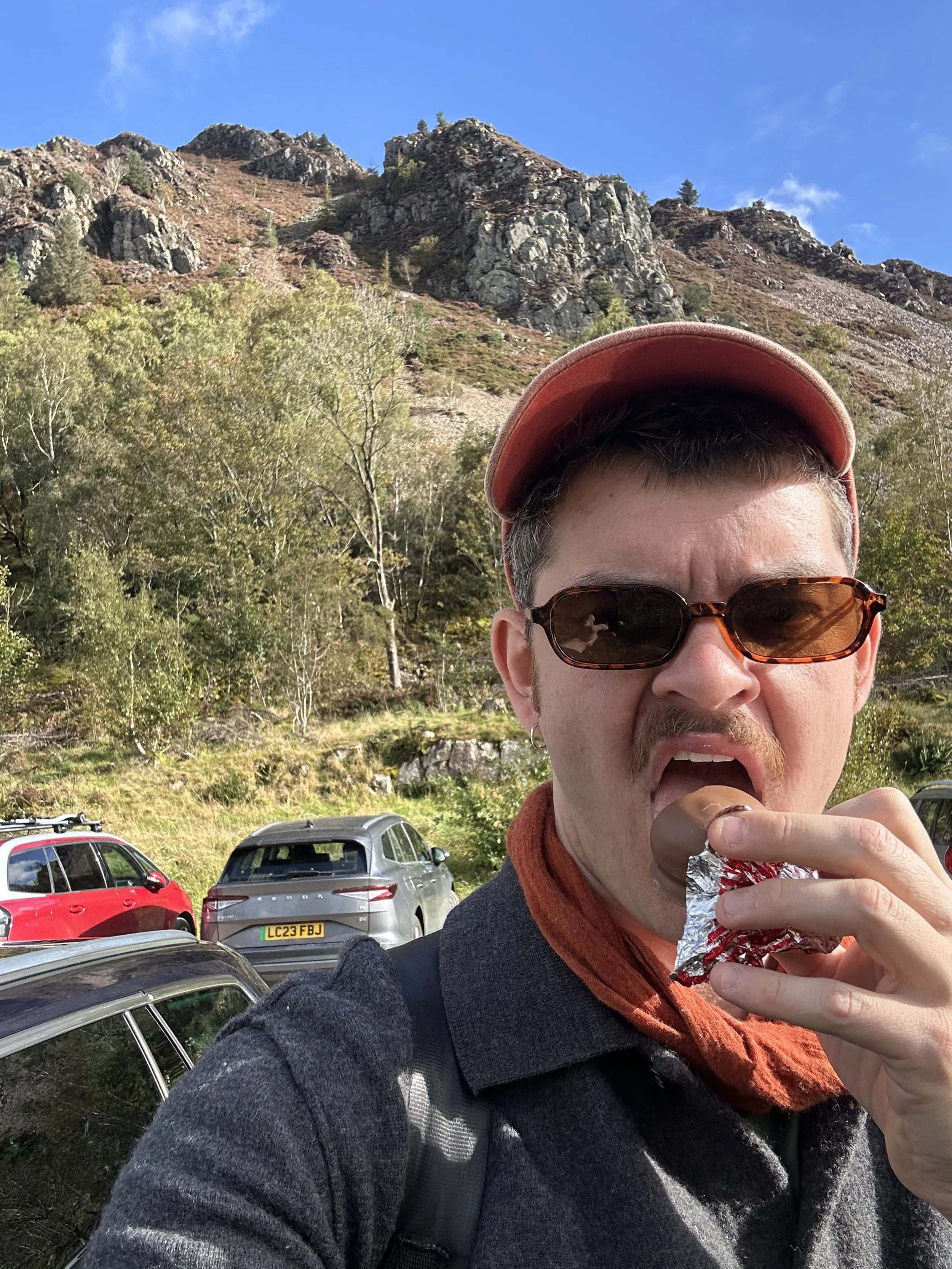 A man wearing sunglasses, a baseball cap, and a scarf eating a chocolate candy bar wrapped in foil outdoors in front of a mountain landscape with cars parked nearby.