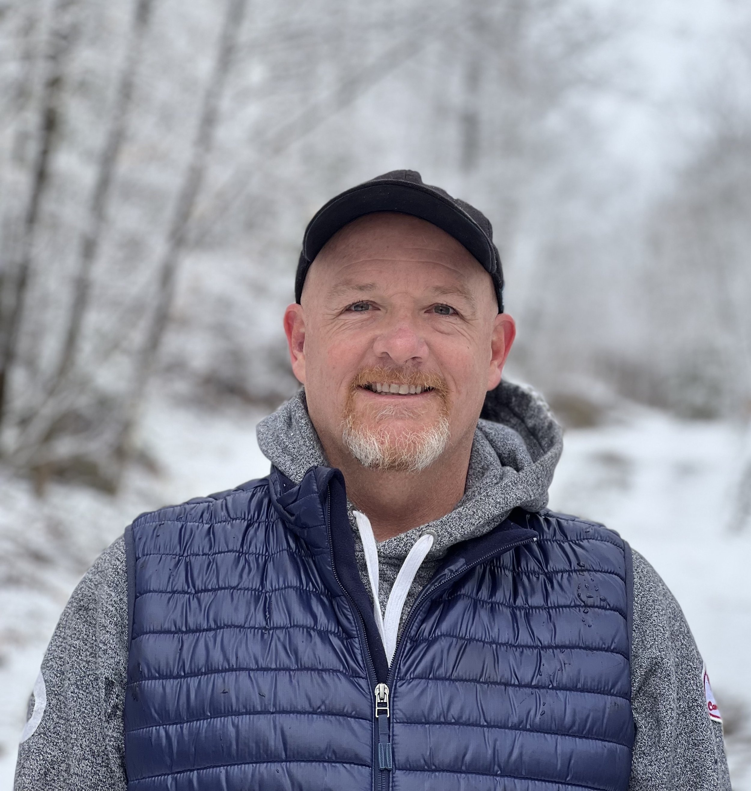 A smiling man wearing a black baseball cap, a gray hoodie, and a navy blue quilted vest, outdoors in a snowy winter landscape.