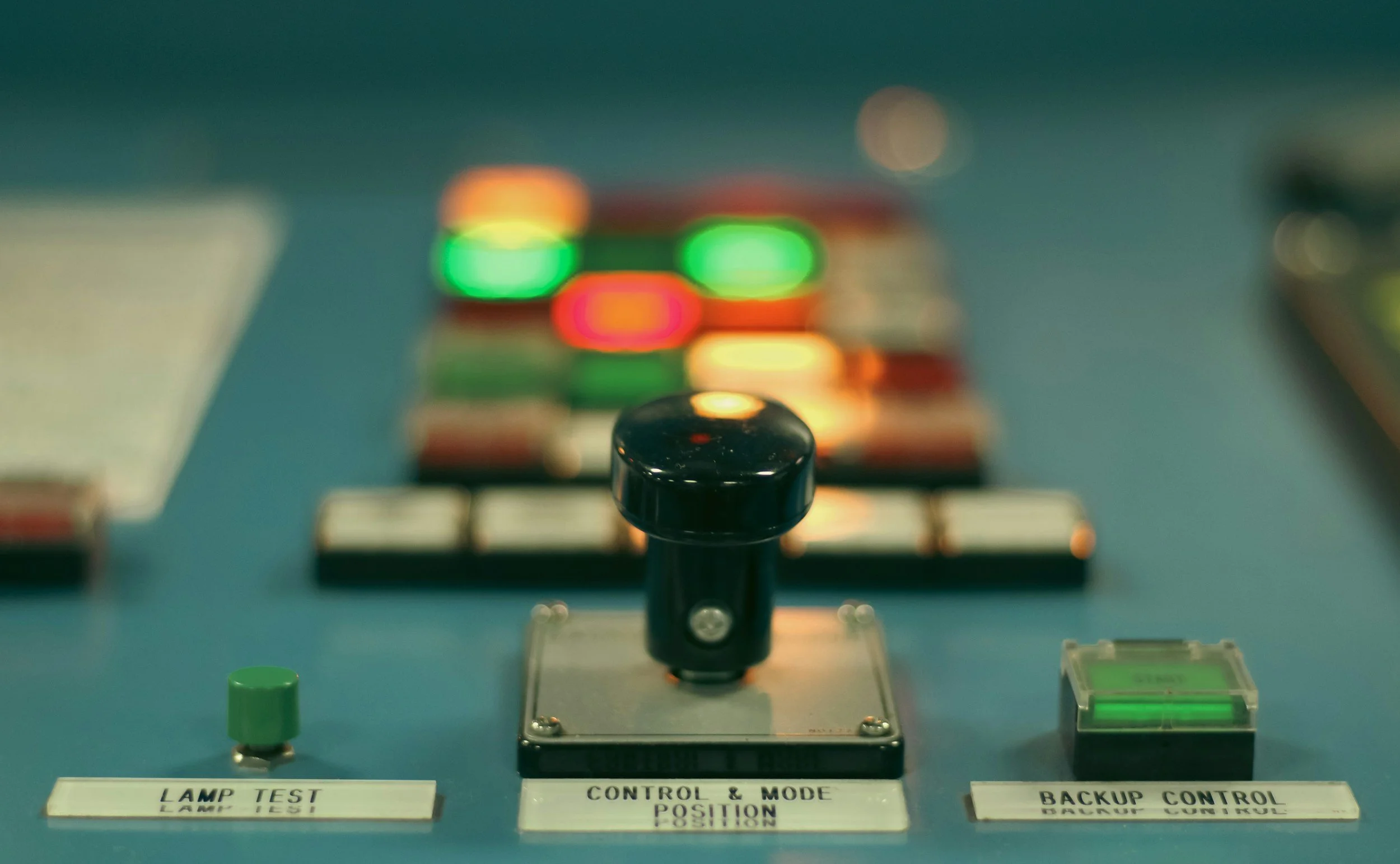 Close-up of control panel with push buttons and switches, including a green lamp test button, a control and mode position switch, and a backup control green indicator, on a blue surface.