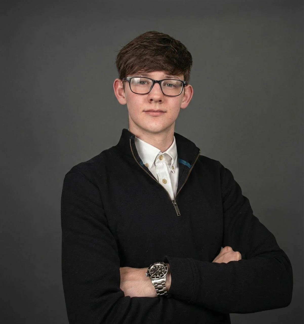 Young man with brown hair and glasses, wearing a white collared shirt under a black zip-up sweater, standing against a gray background with arms crossed, wearing a silver wristwatch.