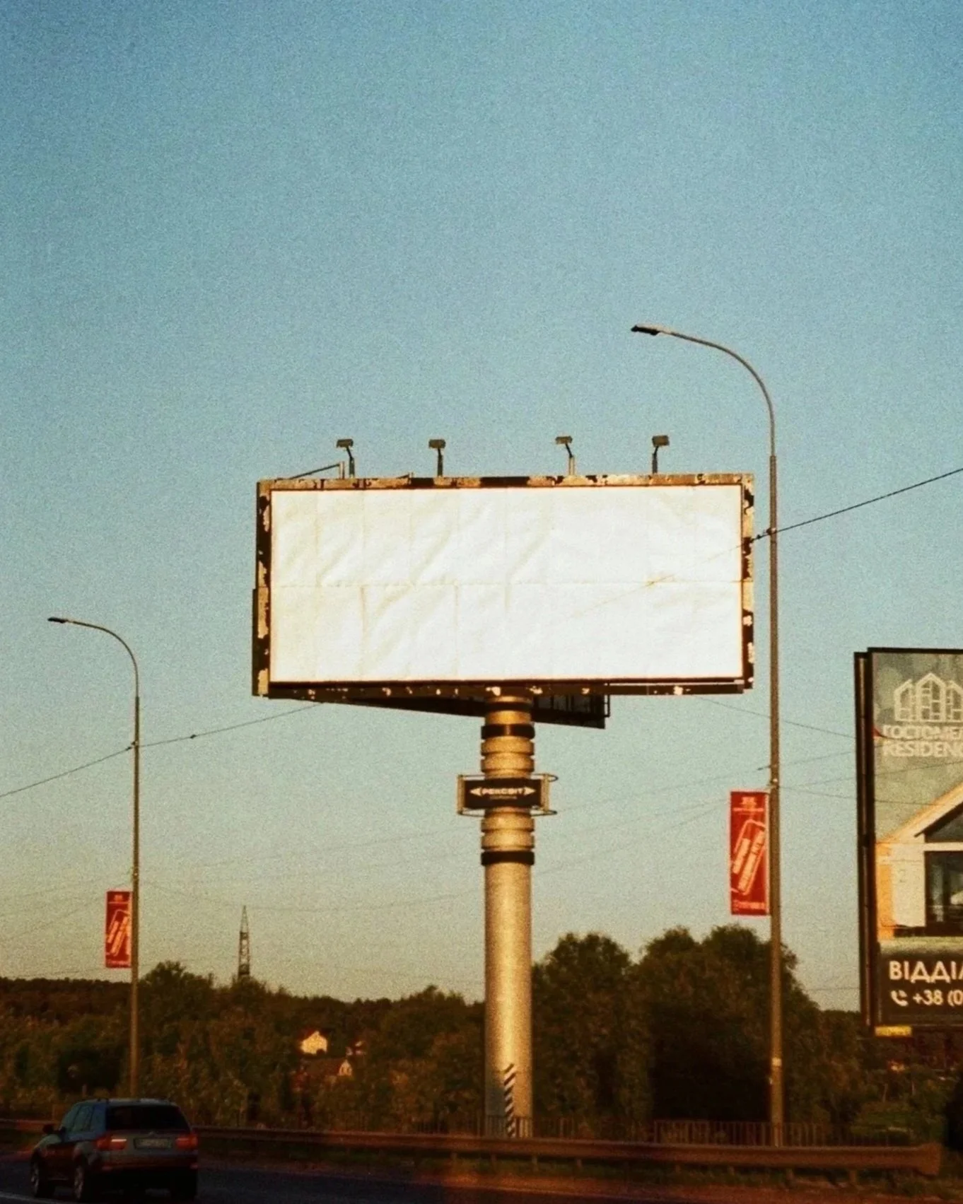 Blank white billboard on a highway with a blue sky background, surrounded by trees and streetlights.