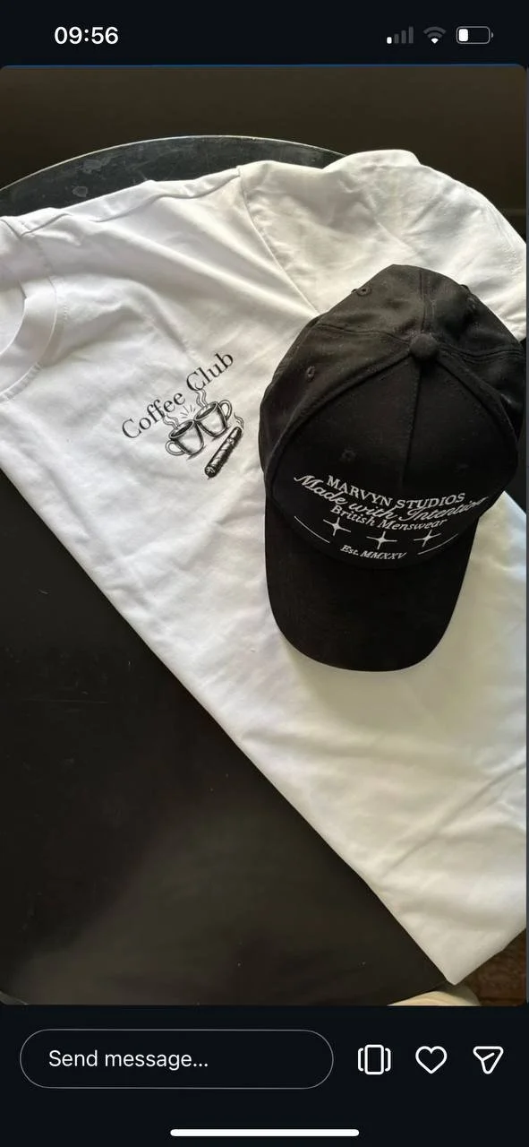 A white t-shirt with a coffee club logo and a black cap with the text 'Marvyn Studios' and 'Made with Otaku' placed on a black surface.