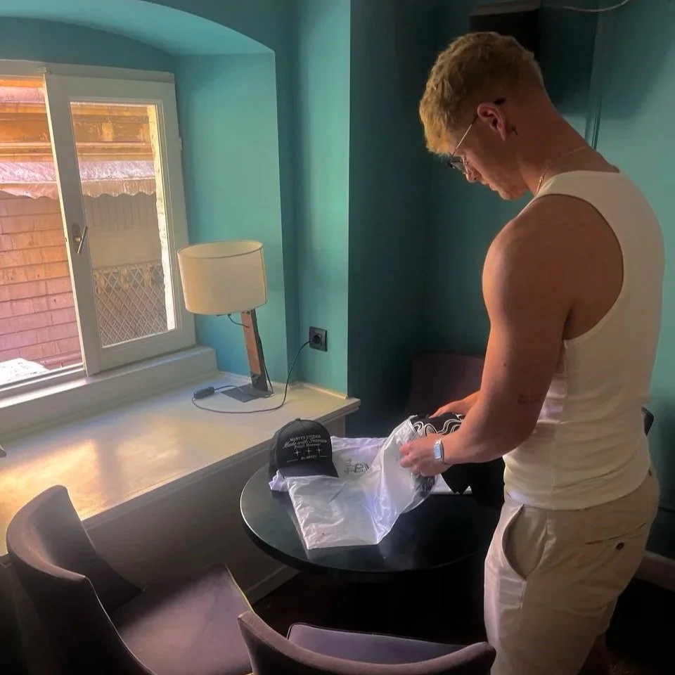 A young man with blonde hair, glasses, wearing a white tank top and beige shorts, unpacking a black cap from a plastic bag in a room with blue walls, near a window with a wooden exterior visible outside.