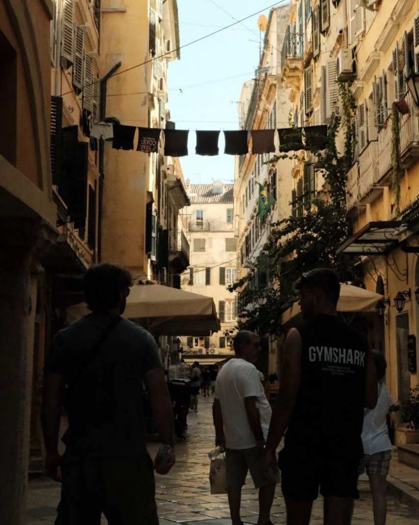 People walking along a cobblestone street lined with multi-story European-style buildings with balconies and shutters, laundry hanging from a line overhead, and umbrellas shading outdoor seating.
