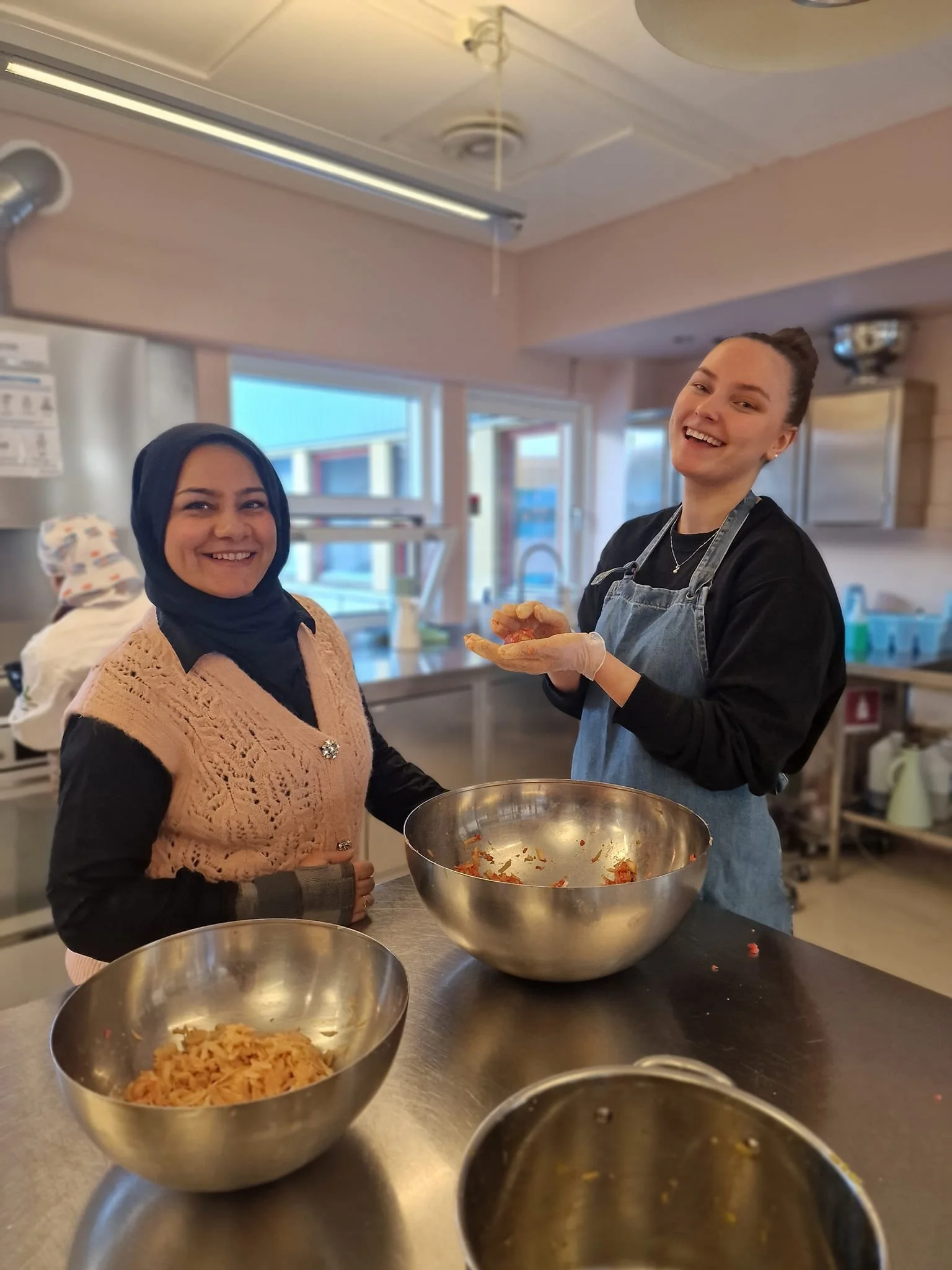 To kvinner lager mat sammen på et kjøkken, én smiler mot kamera, den andre har en skål med ferdigrett, og det er tomme blandeskåler på bordet.